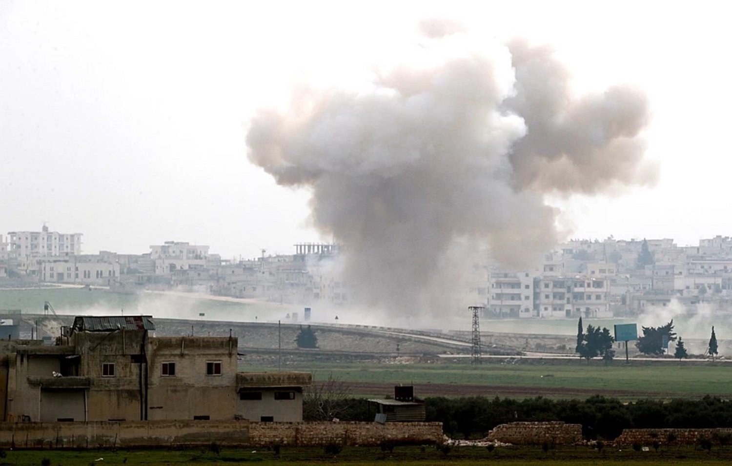 تصویر یک صحنه جنگی را نشان میدهد که در آن دود و گرد و غبار ناشی از یک انفجار دیده میشود. در پسزمینه، ساختمانهای شهری و یک منطقه باز مشاهده میشود. آسمان ابری است و حالت کلی تصویر به موقعیتی پر تنش و خطرناک اشاره دارد. دود در حال بلند شدن از زمین به سمت آسمان است و نشانههایی از ویرانی و نبرد در تصویر مشهود است.