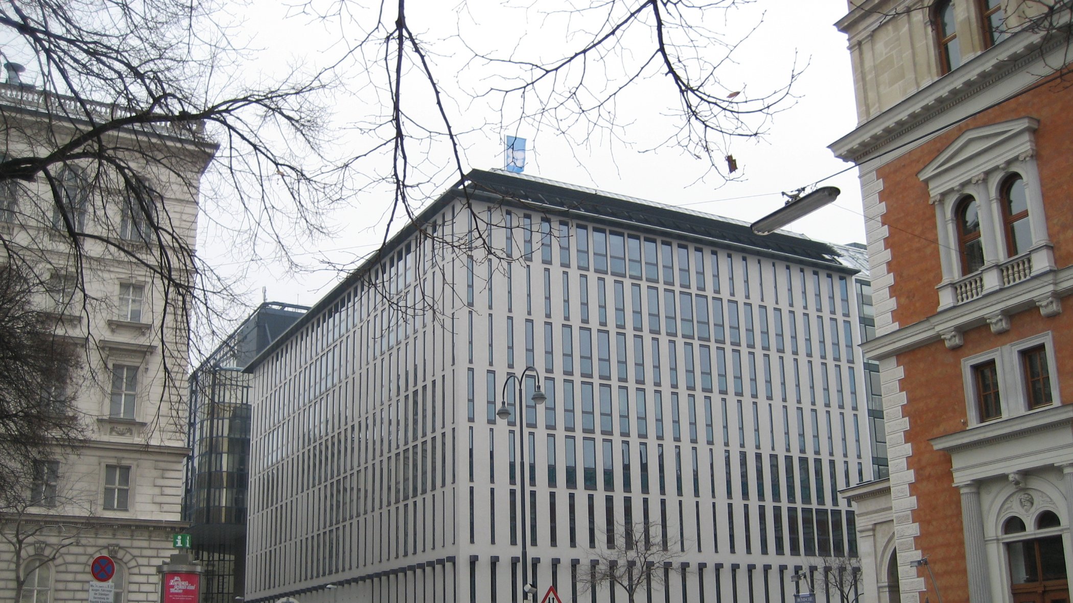 این تصویر نمای یک ساختمان مدرن را نشان میدهد که دارای طراحی مینیمالیستی است. نمای ساختمان با پنجرههای عمودی و خطوط باریک تقسیم شده، ظاهری مرتب و شیک دارد. اطراف ساختمان ممکن است شامل خودروها و درختان بدون برگ در فصل زمستان باشد. در سمت چپ و راست تصویر، ساختمانهای دیگری با طراحیهای کلاسیکتر وجود دارند که تضادی جالب با ساختمان مدرن ایجاد میکنند. فضا به نظر میرسد که در یک منطقه شهری و ممکن است در فصل سرد سال باشد.