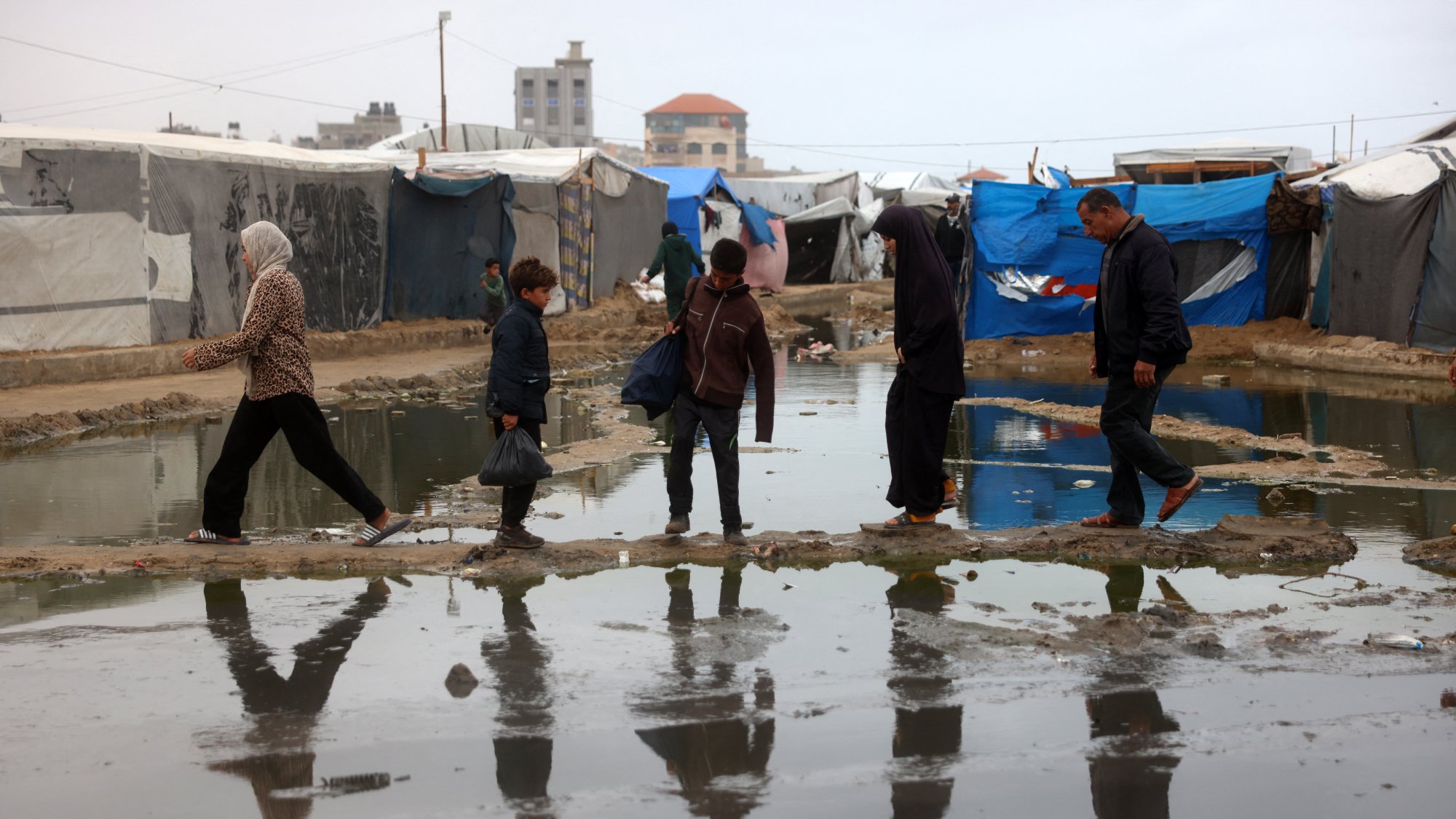 در تصویر، گروهی از مردم در یک منطقه نامناسب عبور میکنند. زیر پاهای آنها آب جمع شده و انعکاس آنها در آب دیده میشود. چادرهای موقتی در زمینه وجود دارند که به وضوح مشخص است. آسمان ابری و فضای کلی تصویر حاکی از شرایط سخت و نامساعد است. افراد در حال حرکت به سمت مقصدی مشخص هستند و لباسهای آنها نشاندهنده وضعیت اقتصادی و اجتماعیاشان است.