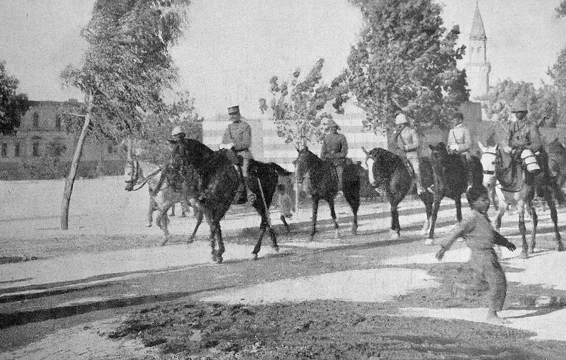 در این تصویر، تعدادی سوارکار باuniform نظامی در حال عبور از یک خیابان هستند. آنها بر روی اسبهای خود سوارند و به سمت جلو حرکت میکنند. در کنار خیابان، یک مرد پیاده در حال عبور است و به سمت دیگر میرود. در پسزمینه، ساختمانهایی دیده میشوند که شاید مربوط به دوره تاریخی خاصی باشند و همچنین یک مناره یا برج در فاصلهای دور قرار دارد. درختان اطراف نیز به تصویر حال و هوای زمان قدیم را میدهند.