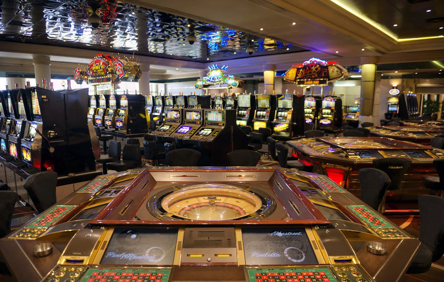 تصویر یک کازینو را نشان میدهد که در آن میزهای قمار و دستگاههای اسلات قرار دارند. در وسط تصویر، میز رولت با چرخ و دکمههای مختلف دیده میشود. در پسزمینه، دستگاههای اسلات و دیگر میزهای قمار به صورت منظم چیده شدهاند. نورپردازی کازینو جذاب و رنگارنگ است و فضای عمومی، نوعی شلوغی و هیجان را به وجود آورده است.
