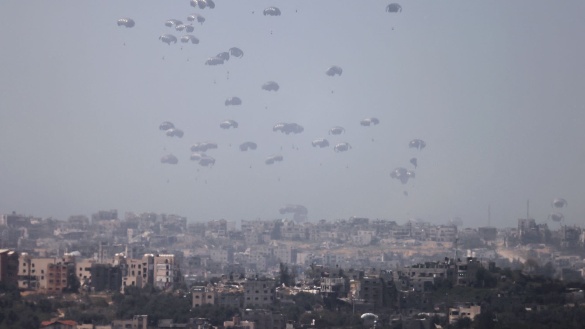 در این تصویر، چندین چتر نجات در آسمان به طور همزمان دیده میشوند که به آرامی در حال سقوط هستند. زمینه تصویر شهری است که شامل ساختمانها و محلههای مختلف است. آسمان ابری و کمی تاریک به نظر میرسد و این باعث میشود فضای تصویر حالتی جدی و نگرانکننده داشته باشد. تصویر به خوبی نمایانگر یک وضعیت نظامی یا اضطراری است.