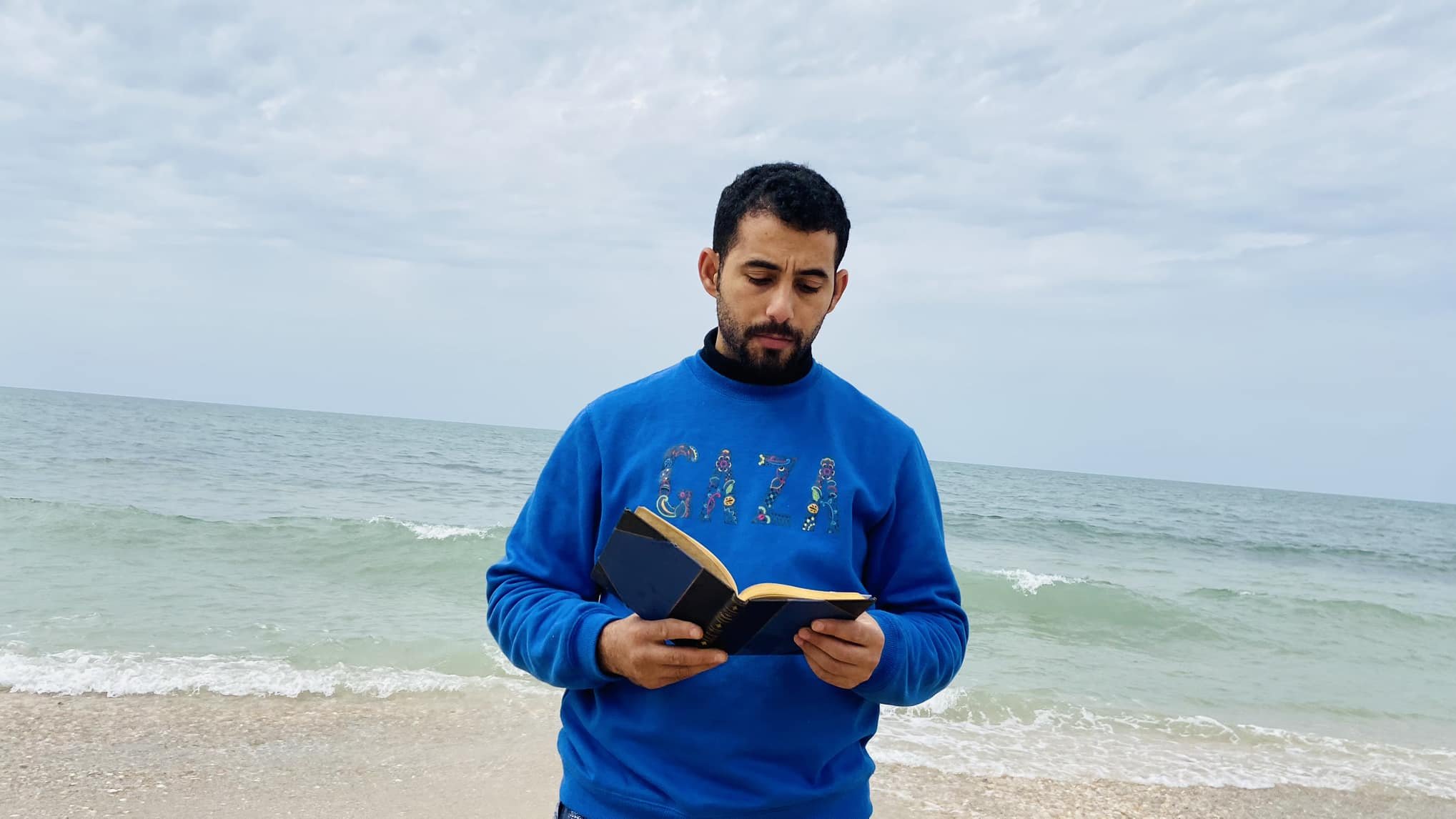 در این تصویر، مردی در کنار دریا ایستاده است و یک کتاب در دست دارد. او لباسی آبی رنگ به تن دارد که روی آن کلمه "GAZA" به چشم میخورد. پسزمینه تصویر را دریای آرام و آسمانی ابری تشکیل میدهد. موجهای دریا به ساحل میرسند و فضای آرامی را به تصویر بخشیدهاند.