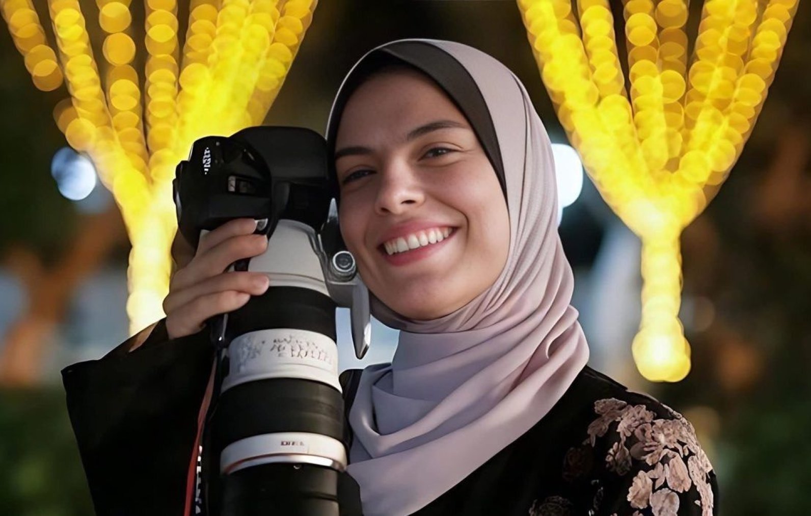 زنی با حجاب، دوربین بزرگ در دست، لبخند بر لب، در پسزمینهای با چراغهای زینتی.