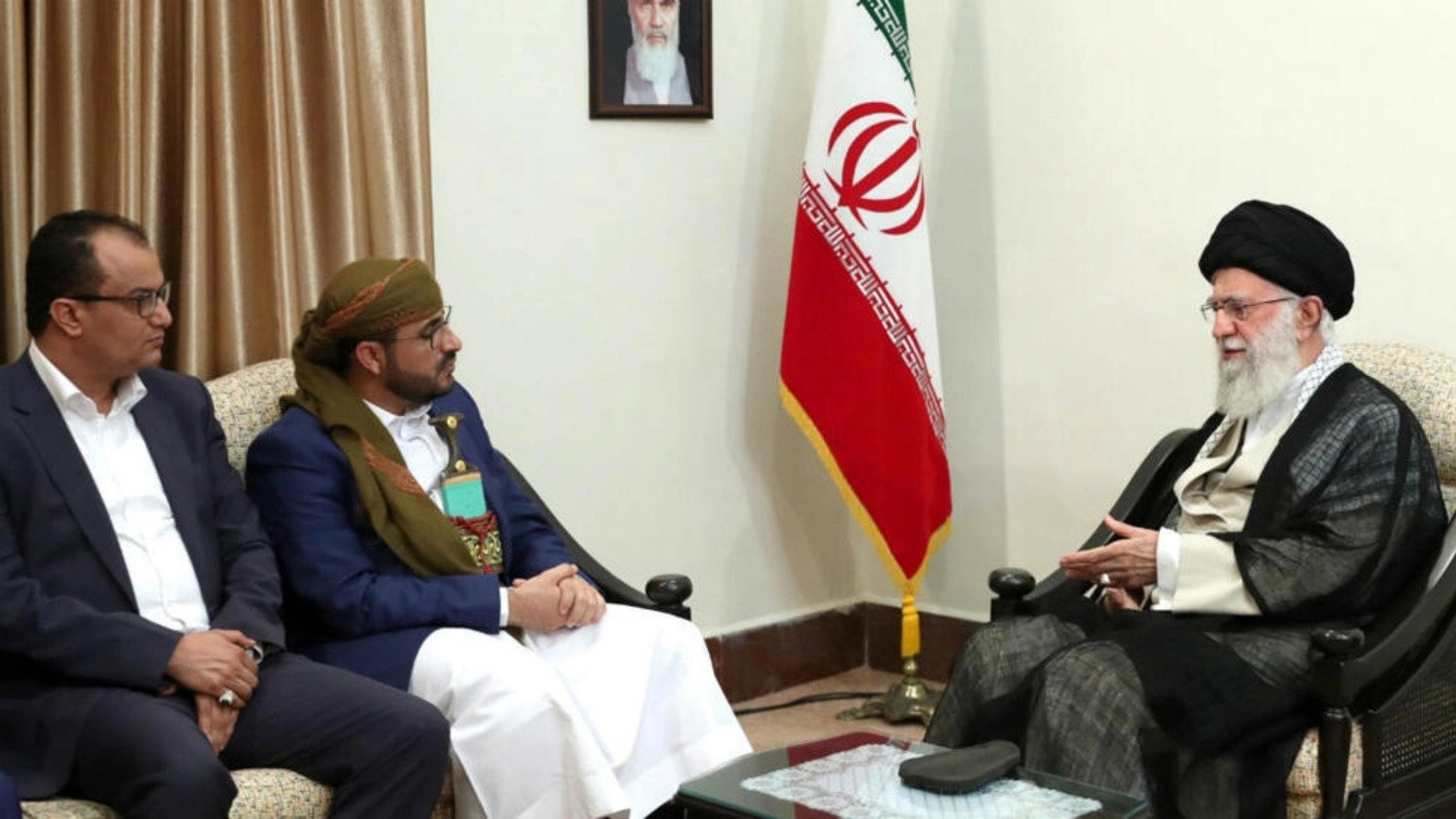 در این تصویر، دو مرد در حال گفتگو با یک شخصیت دیگر هستند. آنها در یک مکان رسمی نشستهاند و تصاویر و پرچمهایی در پسزمینه دیده میشود. یکی از مردان لباس سنتی پوشیده و دیگری لباس رسمی دارد. شخصیت دیگر که صحبت میکند، عمامه و لباس سنتی ایران را به تن دارد. فضای گفتگو نشاندهنده یک جلسه ملاقات رسمی است.