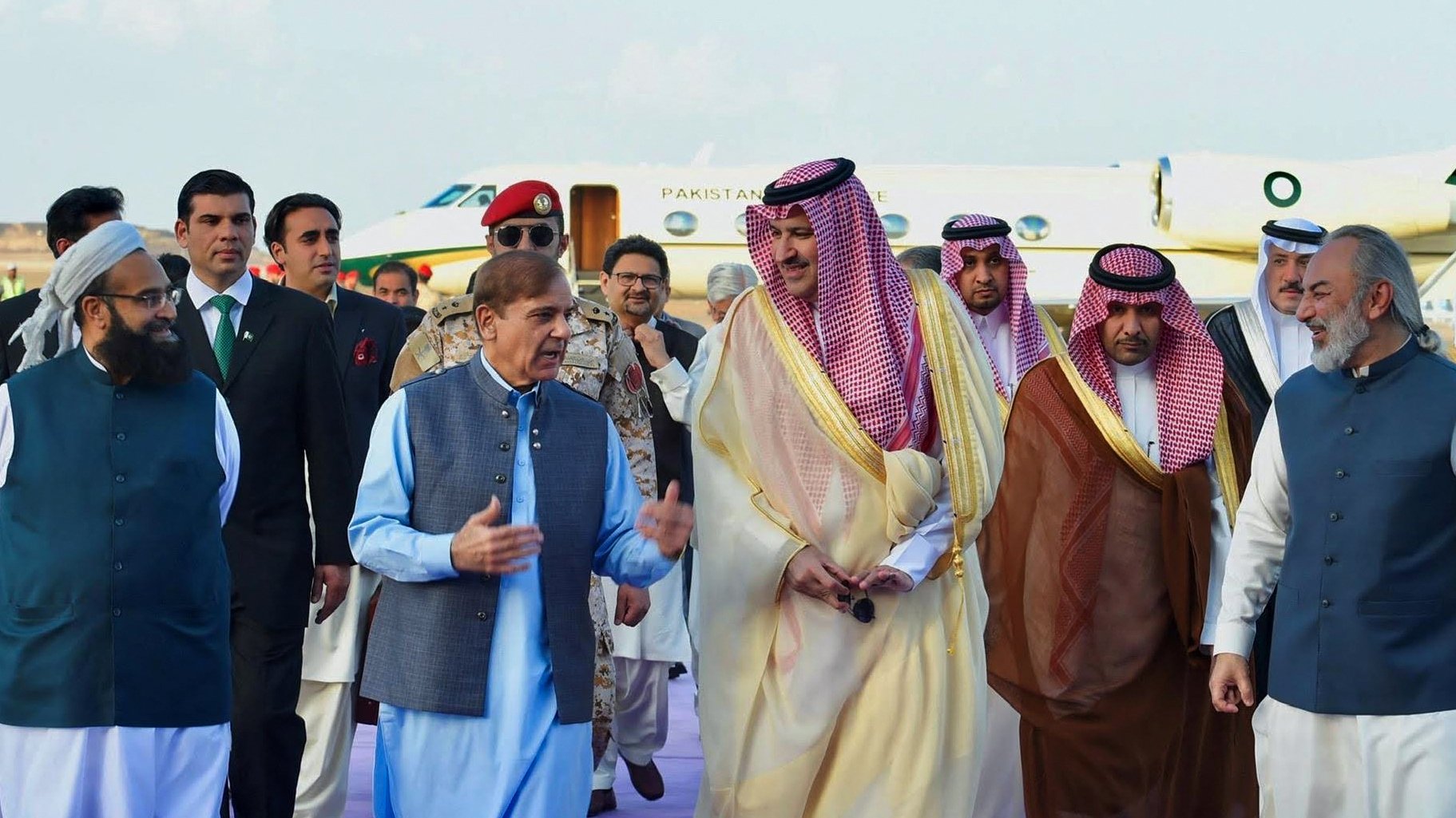 در این تصویر، گروهی از افراد در یک مراسم رسمی یا استقبال دیده میشوند. در مرکز تصویر، مردی با لباس سنتی پاکستانی در حال صحبت با دیگران است. اطراف او افرادی با لباسهای مختلف، از جمله لباسهای رسمی و سنتی، به چشم میخورند. پسزمینه شامل یک هواپیمای خصوصی است که نشاندهنده موقعیت ویژه این ملاقات است. فضا به نظر میرسد که محترمانه و دوستانه باشد.