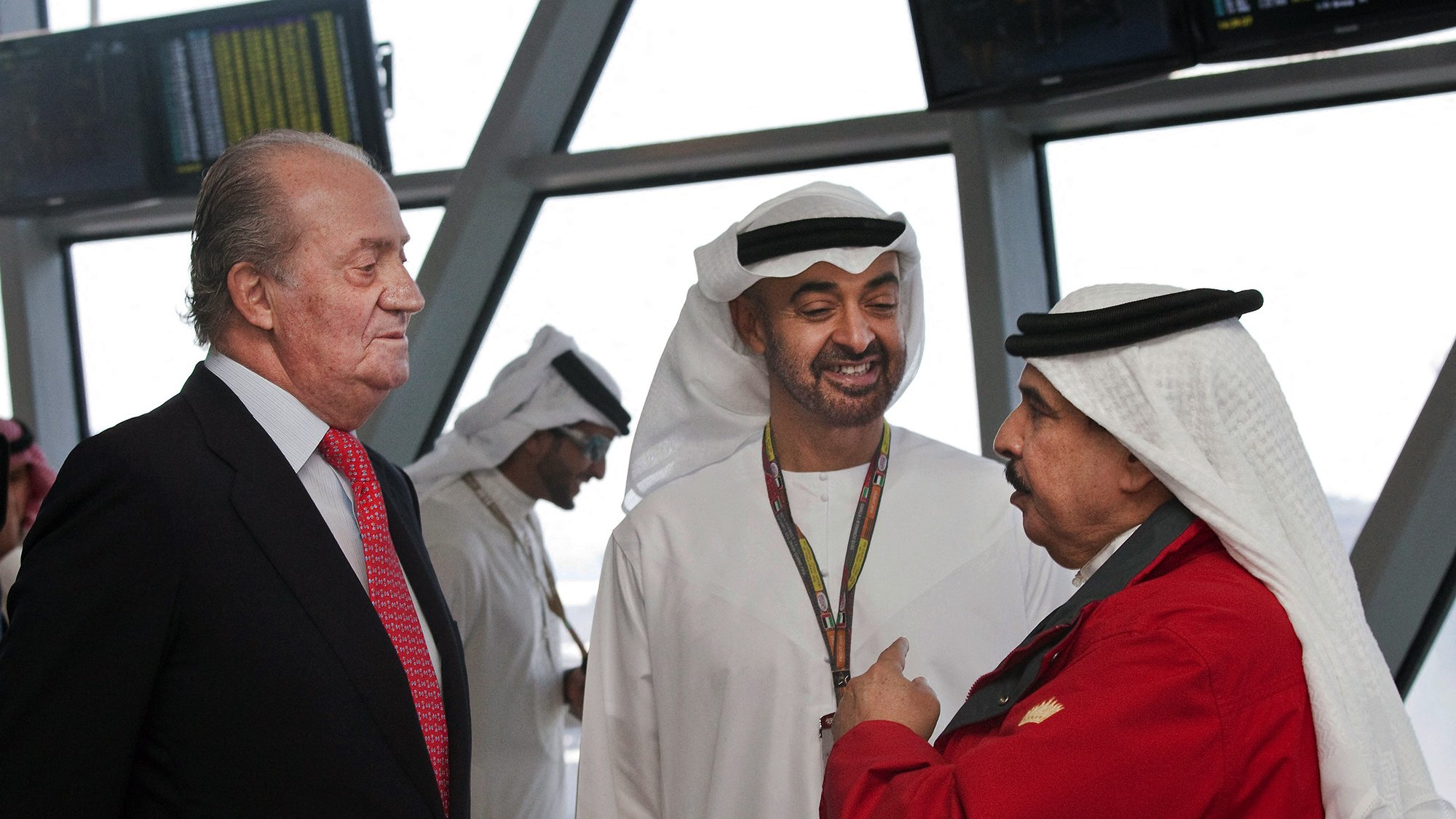 در این تصویر، سه مرد در یک مکان رسمی ایستادهاند. یکی از آنها که لباس رسمی پوشیده، به نظر میرسد که از چهرهای جدی برخوردار است. دو نفر دیگر لباس سنتی عربی به تن دارند. یکی از آنها لباسی سفید و دیگری لباسی قرمز به همراه کلاه عربی دارد. آنها به طور دوستانه با یکدیگر صحبت میکنند و در پسزمینه میتوان تعدادی دیگر از افراد را دید که در حال حرکت هستند. فضای تصویر نشاندهنده یک مکان شلوغ و مدرن است.