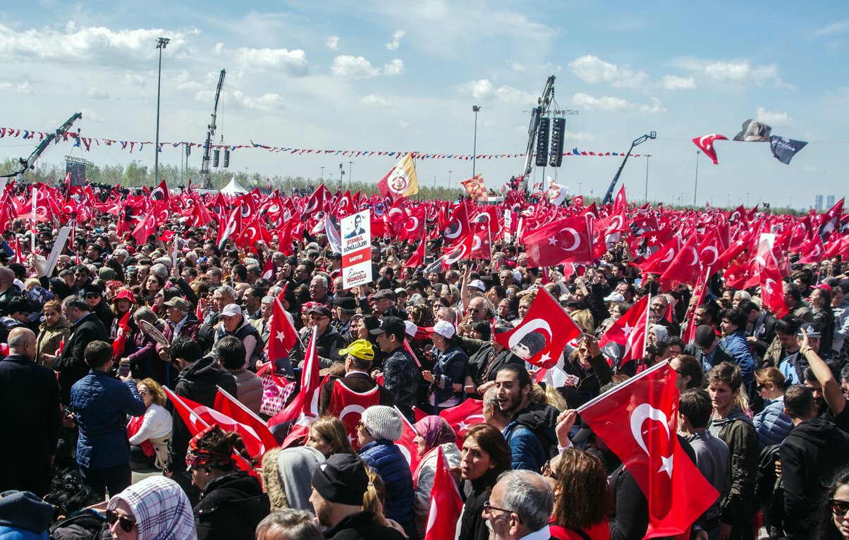 تصویر نشاندهنده تظاهرات بزرگی در یک فضای باز است که تعداد زیادی از مردم حضور دارند. آنها پرچمهای قرمز و سفید ترکیه را در دست دارند و به نظر میرسد که در حال ابراز همبستگی یا حمایت از یک حرکت یا رویداد خاص هستند. آسمان آبی است و چند ابر سفید در آن دیده میشود. جمعیت متنوعی در تصویر وجود دارد که نشاندهنده حضور افراد با سنین و زمینههای مختلف اجتماعی است. فعالیت و شور و حال در این جمعیت به وضوح قابل مشاهده است.