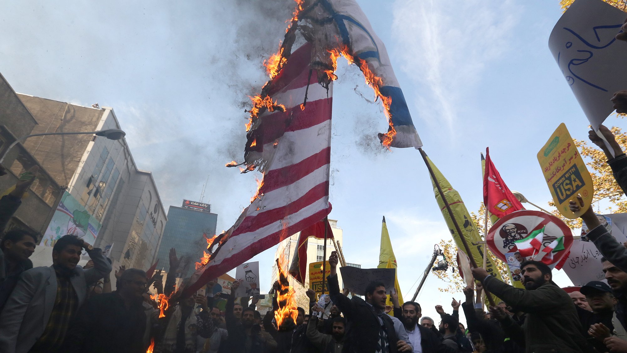 در این تصویر، جمعیتی در حال برگزاری یک تظاهرات هستند. گروهی از افراد در حال آتش زدن پرچمهایی با نمادهای مشخص هستند. پشت زمینه تظاهرات، ساختمانها و تابلوهای تبلیغاتی دیده میشود. شمار زیادی از تظاهرکنندگان در دست خود پلاکاردهایی نشان میدهند که بر روی آنها شعارهایی نوشته شده است. جو عمومی تصویر نشاندهندهٔ هیجان و احساسات قوی شرکتکنندگان است.