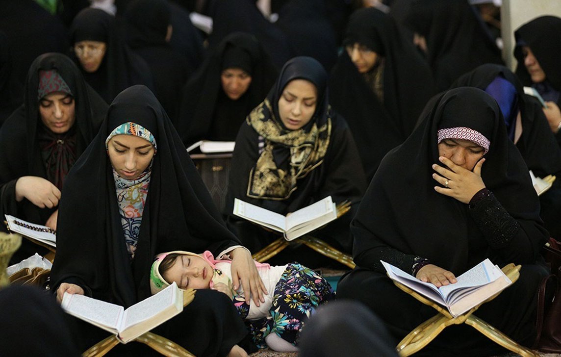در تصویر گروهی از زنان با چادرهای سیاه در حال مطالعه قرآن دیده میشوند. بعضی از آنها در حال خواندن هستند و توجه عمیقی به کتاب دارند. همچنین، یک کودک در آغوش یکی از زنان خوابیده است. بهطور کلی، فضایی معنوی و پر از تمرکز در این تصویر وجود دارد.