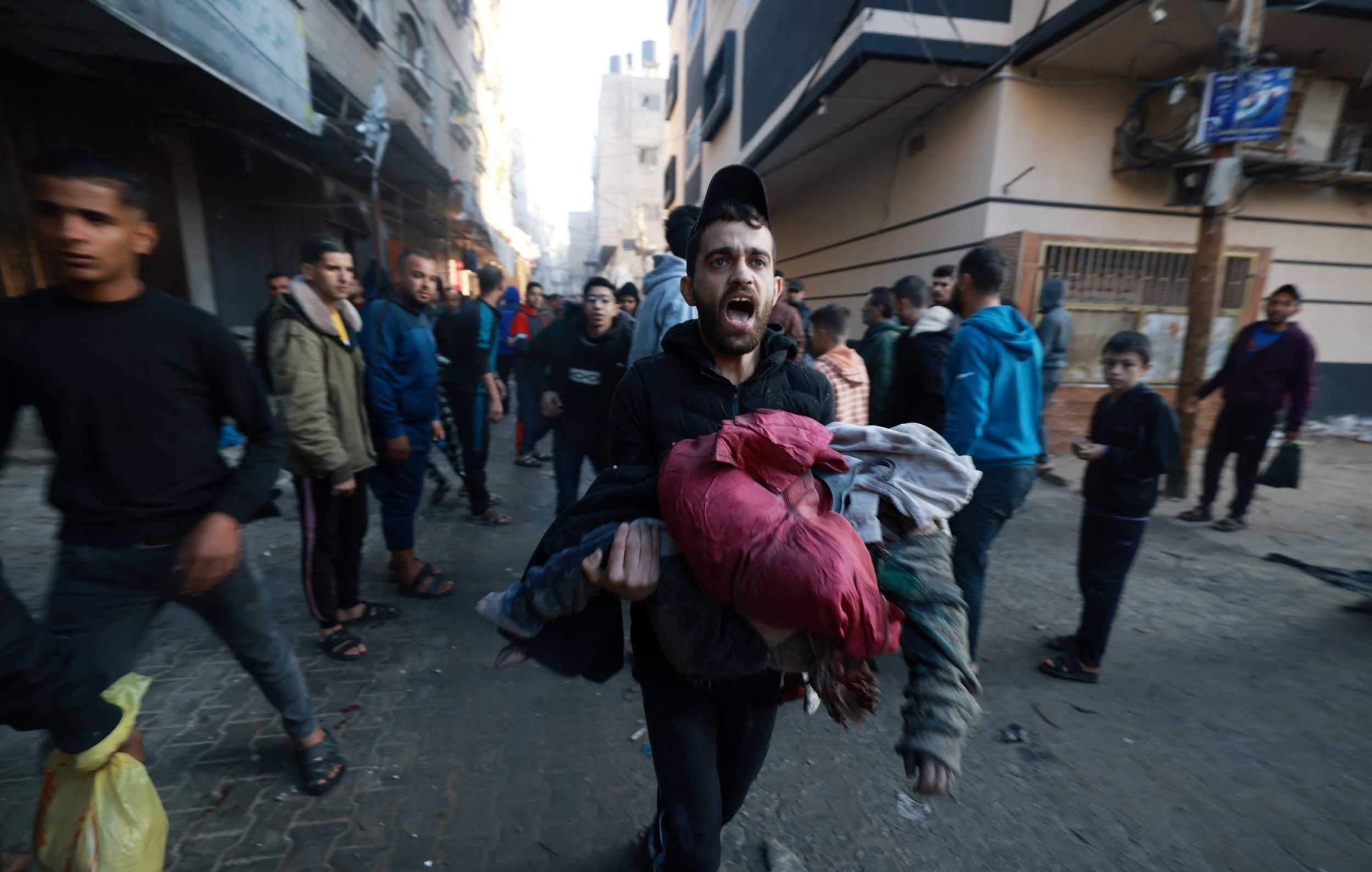 در این تصویر، گروهی از مردم در یک خیابان مشغول حرکت هستند، به نظر میرسد که موقعیت اضطراری یا بحرانی وجود دارد. یک مرد در حال دویدن است و چندین لباس را در آغوش گرفته است. چهره او حاکی از اضطراب و نگرانی است. دیگر افراد نیز در حال حرکت هستند و فضای خیابان به نظر شلوغ و پر از فعالیت است. ساختمانها در پسزمینه نشاندهنده یک محیط شهری هستند.