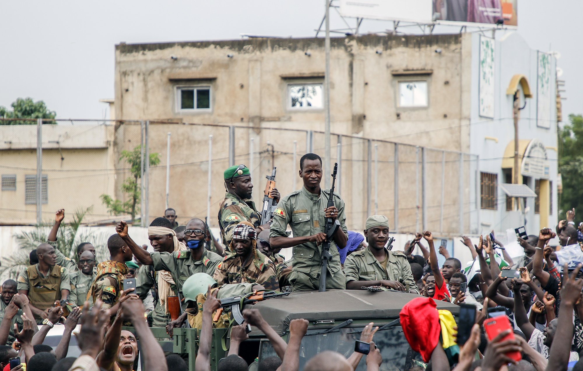 در این تصویر، تعدادی نظامی با لباس نظامی و سلاح در دست در بالای یک خودروی نظامی ایستادهاند. اطراف آنها جمعیتی از مردم دیده میشود که در حال خوشحالی و حمایت از نظامیها هستند و دستهایشان را بالا بردهاند.背景 تصویر نیز ساختمانهایی دیده میشود که نشان از یک فضای شهری دارد. احساس این تصویر نشاندهنده شور و هیجان جمعیت به خاطر نظامیها است.