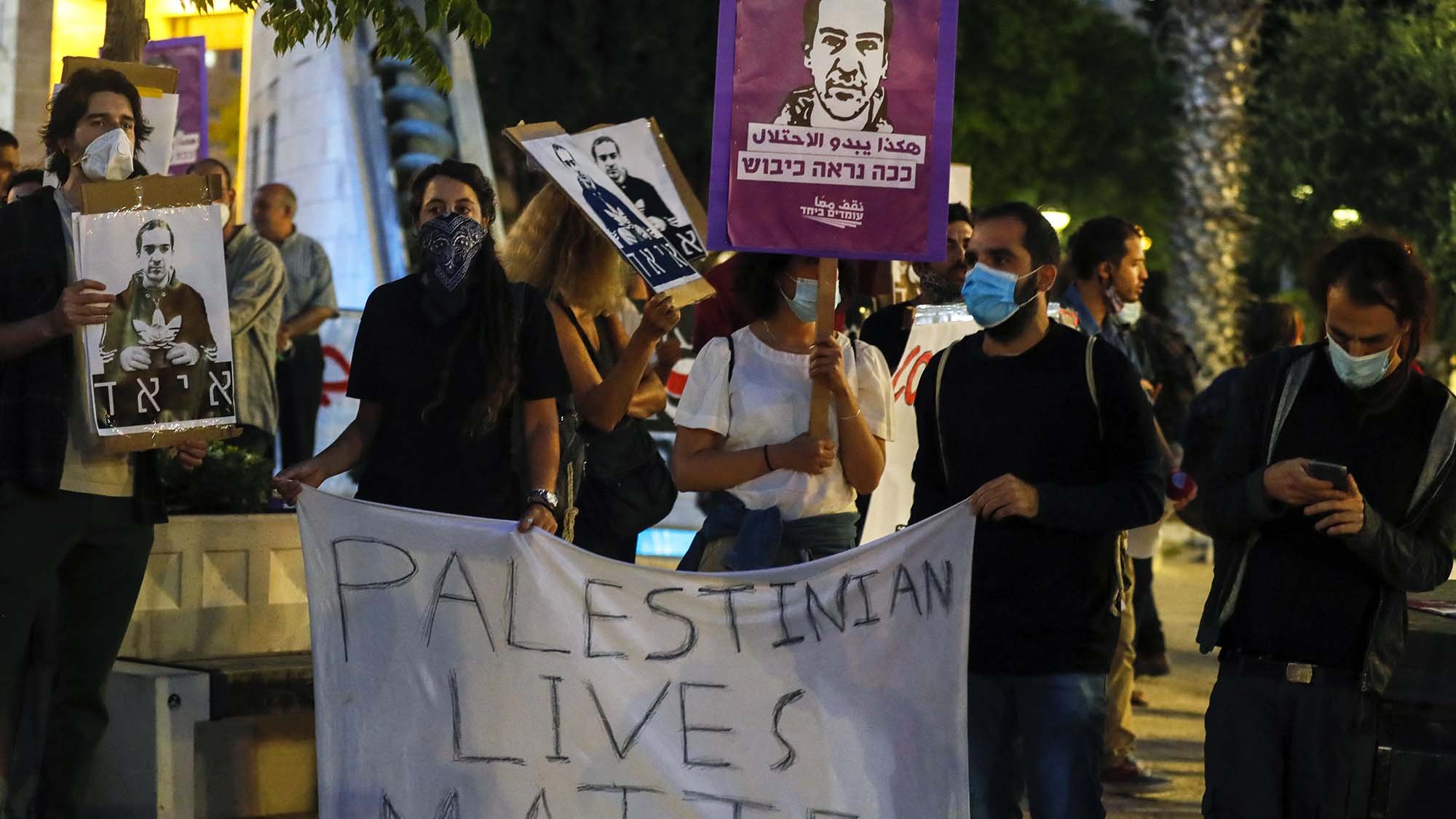 در این تصویر، گروهی از مردم در حال برگزاری یک تجمع اعتراضی هستند. آنها تابلوهایی با تصاویر و شعارهایی در دست دارند. یکی از بنرهای بزرگ به زبان انگلیسی میگوید "زندگیهای فلسطینی مهم است". شرکتکنندگان به وضوح از ماسک استفاده میکنند، و فضای عمومی تجمع نشاندهنده یک اعتراض اجتماعی است. در پسزمینه، درختان و یک محیط شهری قابل مشاهده است.