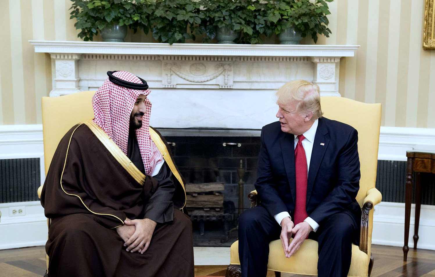 در این تصویر، دو نفر در یک اتاق نشستهاند. یکی از آنها لباس سنتی عربی به تن دارد و از لحاظ ظاهری به عنوان یک مقام عربی به نظر میرسد. فرد دوم، لباس رسمی غربی پوشیده و به نظر میرسد یک مقام سیاسی است. آنها در حال گفتگو هستند و هر دو به همدیگر نگاه میکنند. محیط اتاق با دیوارهای روشن و دکوراسیون رسمی طراحی شده است. در پسزمینه، شومینهای وجود دارد که به فضای اتاق اضافه میکند. گیاهان سبز نیز در بالای شومینه قرار دارند.