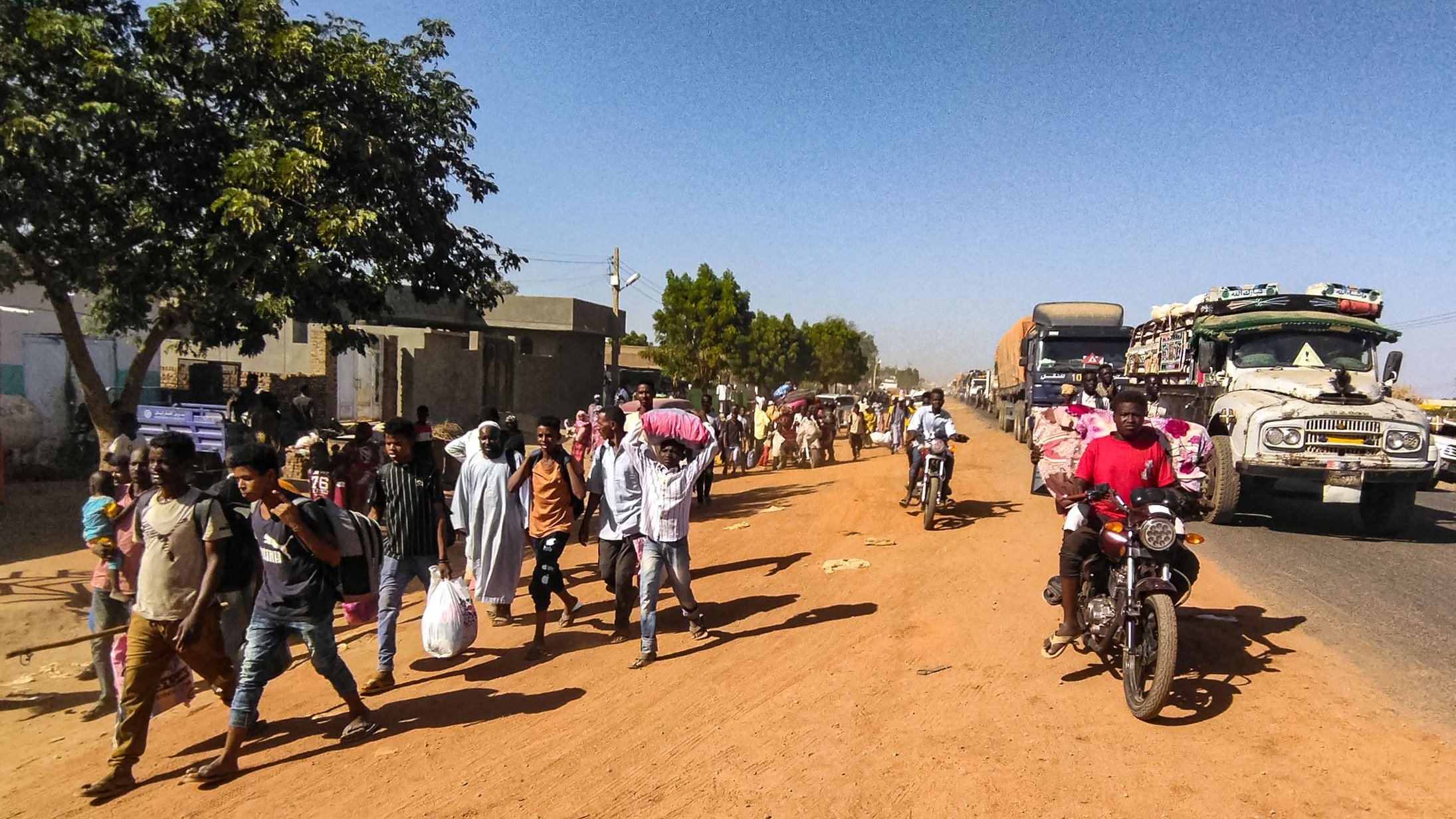 در این تصویر، یک خیابان شنی و پرجنبوجوش دیده میشود. در سمت چپ، تعدادی درخت و ساختمانهای ساده قرار دارند. گروهی از مردم در حال پیادهروی هستند و برخی از آنها کیسههایی به دوش دارند. در سمت راست، چندین خودرو و موتورسیکلت در حال تردد هستند. فضای عمومی پر از فعالیت و زندگی به نظر میرسد.