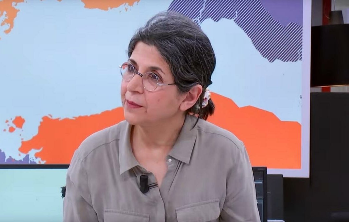 این تصویر فردی را نشان میدهد که در یک محیط خبری یا استودیو قرار دارد. او دارای موهای خاکستری و آرایش ساده است و عینک دارد. لباس او رسمی و به رنگ ملایم است. پسزمینه تصویر نقشهای از جهان یا بخشهایی از آن را نمایش میدهد که به نظر میرسد به موضوع خبر مربوط میشود. چهره این فرد نشاندهنده توجه و تمرکز است.