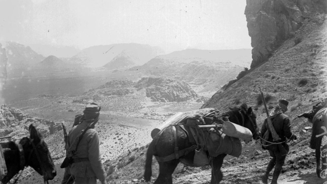 در این تصویر، گروهی از نظامیها در حال عبور از یک مسیر کوهستانی هستند. تعدادی از آنان در حال پیادهروی هستند و برخی دیگر بر روی اسبها یا الاغها سوارند. چشمانداز طبیعی در زمینه شامل کوهها و دشتهای وسیع است. تصویر حال و هوای قدیمی و تاریخی دارد و نمایی از زندگی نظامی در منطقه کوهستانی را به تصویر میکشد.