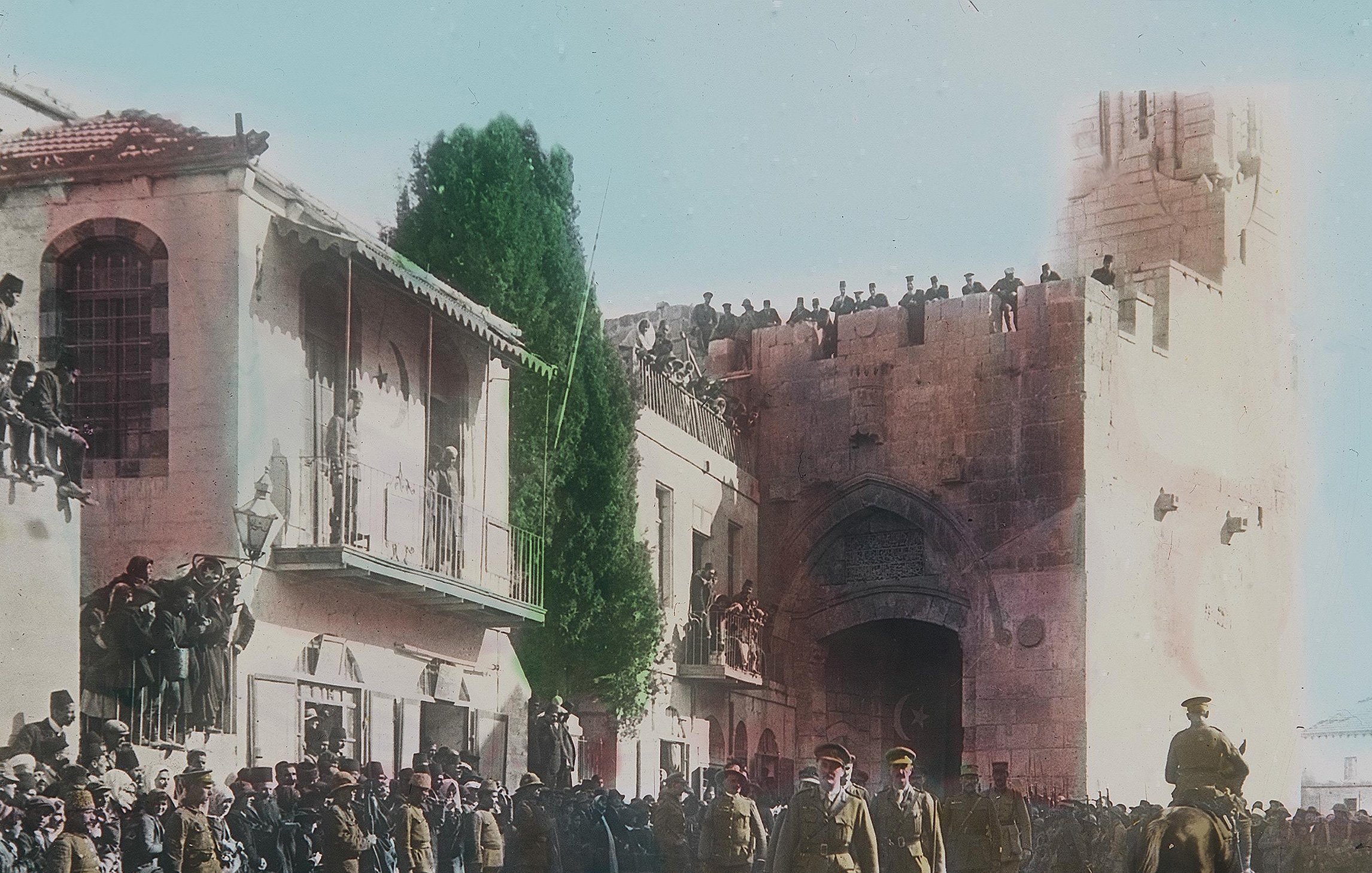 در این تصویر تاریخی، یک گروه از نظامیان در حال عبور از خیابانی در شهری با معماری قدیمی هستند. در دو طرف خیابان، جمعیتی زیادی ایستادهاند و به تماشای اتفاقات میپردازند. در پسزمینه، ساختمانهایی با طراحی خاص و دیواری بزرگ دیده میشوند. برخی از افراد در بالای دیوار ایستادهاند و نظارهگر هستند. فضای تصویر حسی از یک رویداد رسمی یا مراسم ویژه را القا میکند و رنگآمیزی تصویر به آن جذابیت بیشتری بخشیده است.