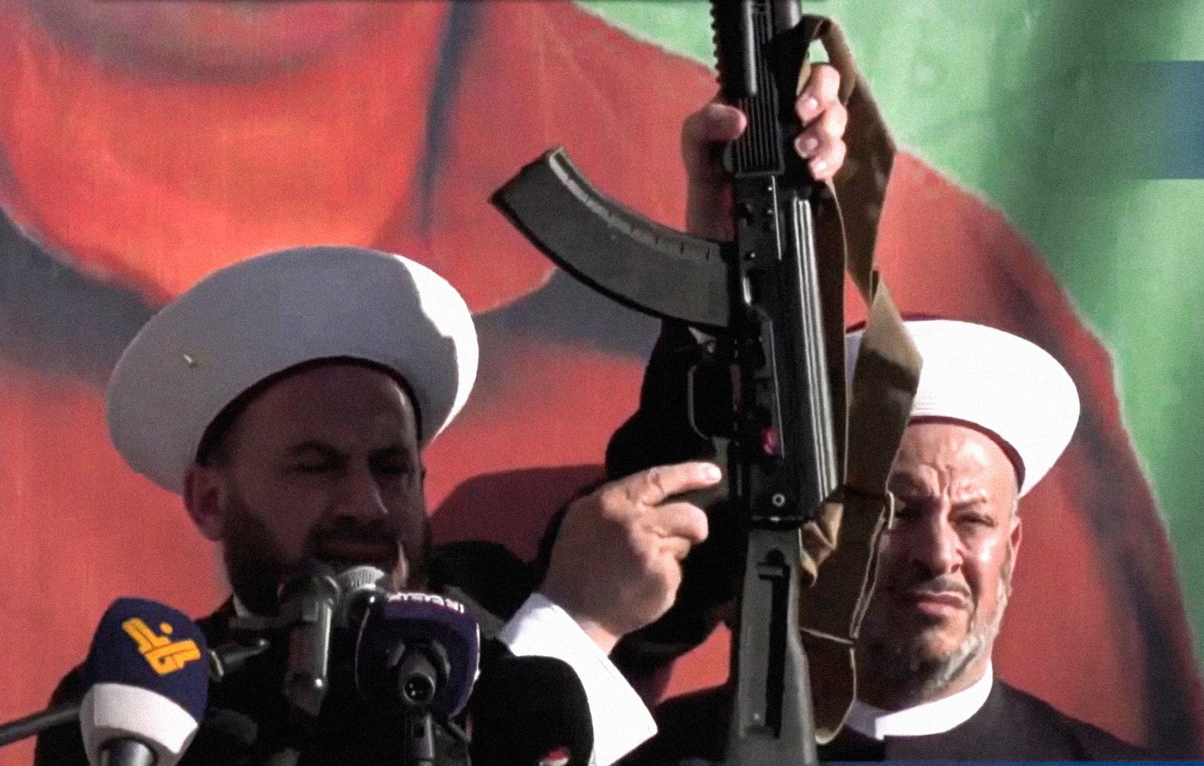 در این تصویر، دو فرد که در حال سخنرانی هستند، به نظر میرسد در یک مراسم یا تجمع حضور دارند. یکی از آنها سلاحی را در دست دارد و دیگر فرد در سمت راست نیز در حال نظارت یا همراهی با او است. پسزمینه تصویر شامل رنگهای زنده و گرافیکی است. هر دو فرد کلاههای مخصوص خود را بر سر دارند که نشاندهنده هویت مذهبی یا فرهنگی آنها است. احساس کلی تصویر به نظر میرسد جدی و تأکیدی بر موضوعی مهم باشد.