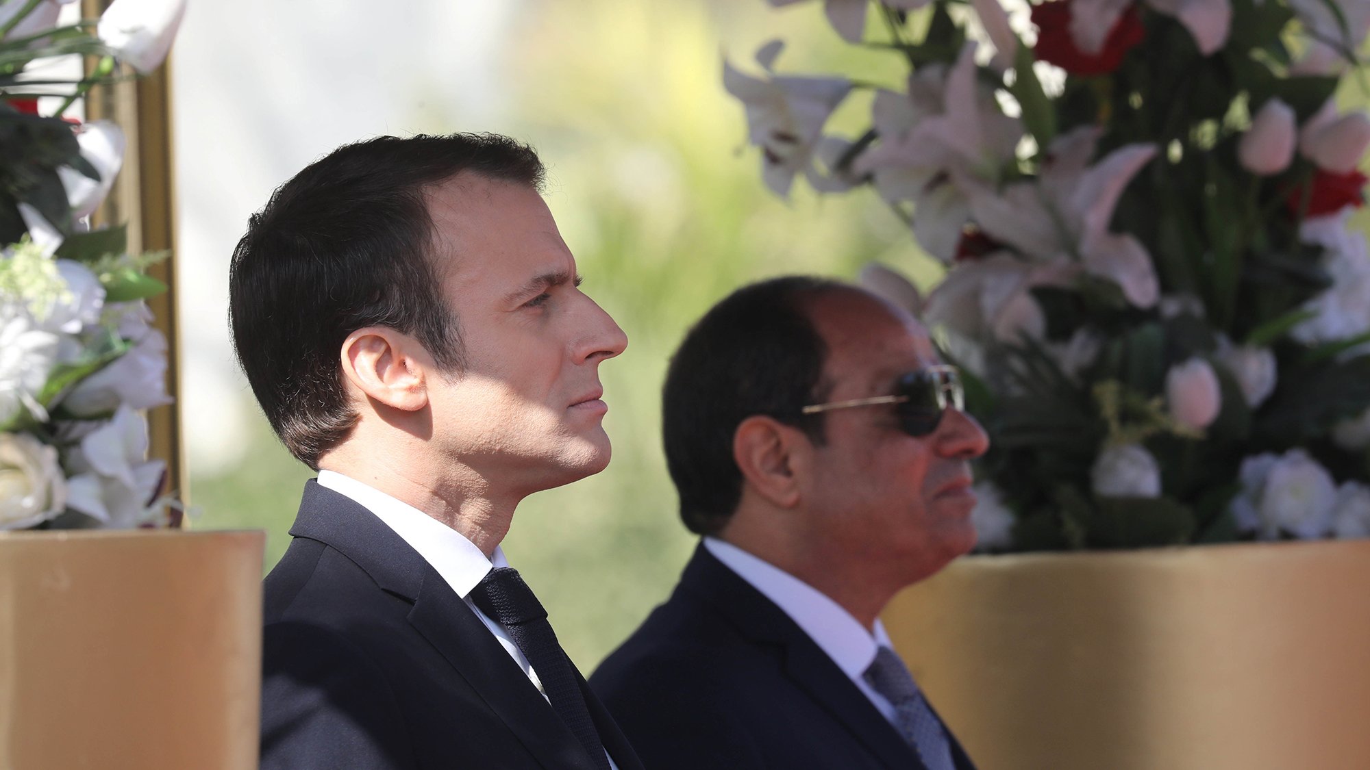 این تصویر دو مرد را در کنار هم نشان میدهد. شخصی در سمت چپ با موهای تیره و لباس رسمی مشکی، در حالت تفکر و جدی ایستاده است. در سمت راست، مرد دیگری با عینک آفتابی و لباس رسمی به رنگ تیره، در کنار او قرار دارد. پسزمینه شامل گلهای سفید و عناصر طبیعی است که به تصویر زیبایی و شکیل بودن میبخشد.