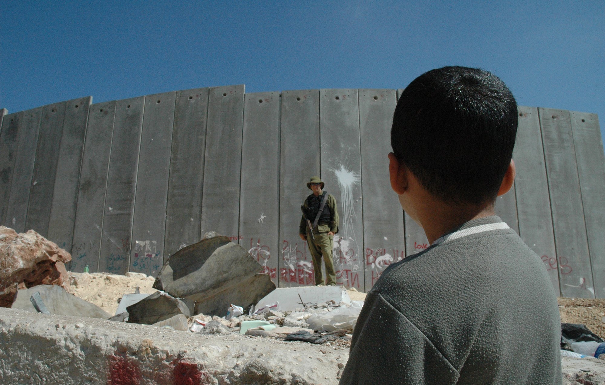 در این تصویر، پسر بچهای به سمت یک دیوار بلند و بتنی نگاه میکند. دیوار دارای آثار graffiti و علامتهای مختلفی است. در مقابل او، یک نظامی ایستاده است که لباس نظامی به تن دارد و به نظر میرسد در حال نظارت است. محیط اطراف شامل سنگها و زبالههای پراکنده است و آسمان آبی و روشنی بالای سر آنها دیده میشود. احساس تصویر میتواند تنش و جدایی را به خوبی نشان دهد.