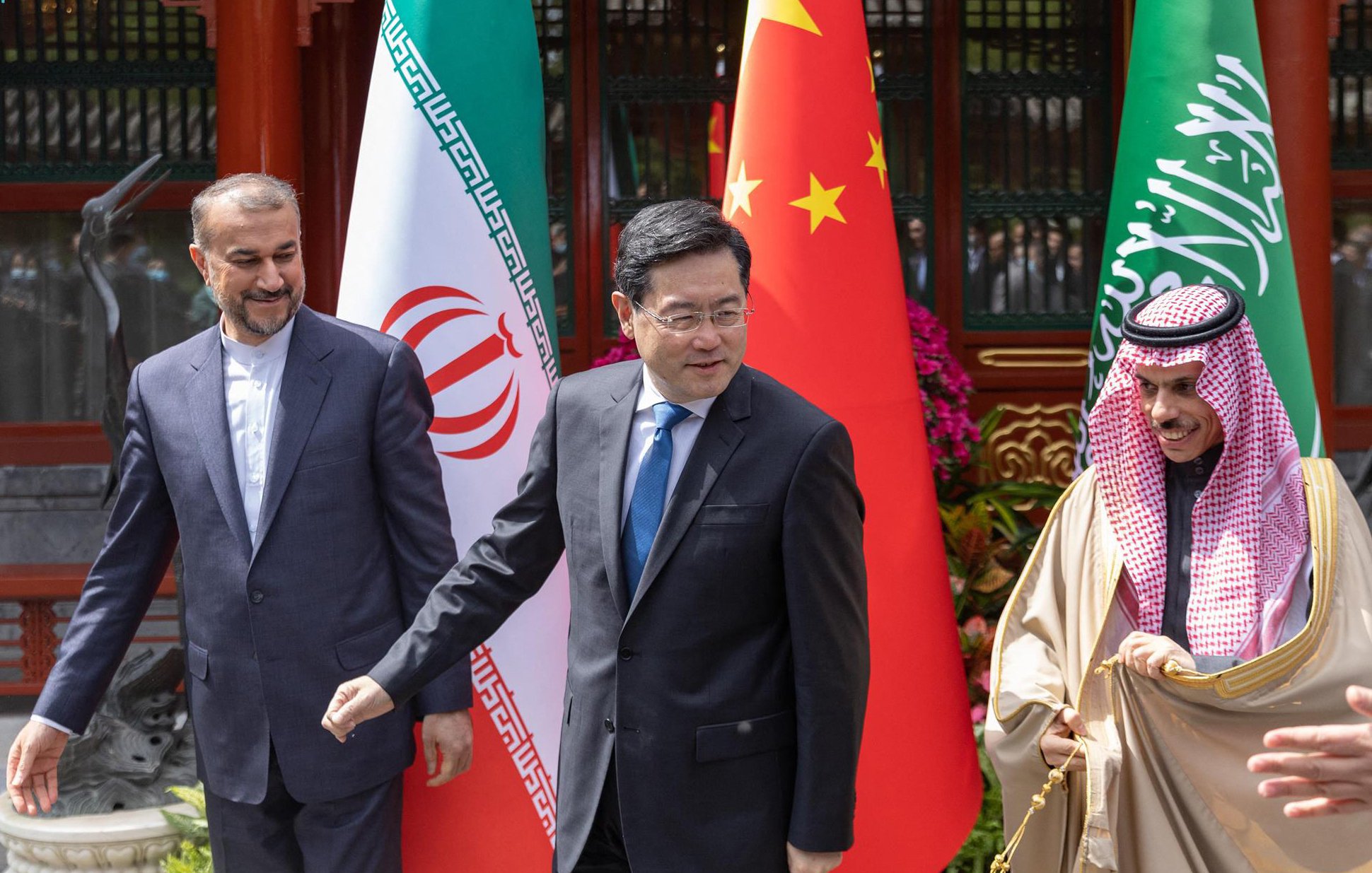 در این تصویر، سه نفر در حال ایستادن و گفتگو در یک محیط رسمی هستند. در سمت چپ، مردی با لباس رسمی و کت و شلوار تیره دیده میشود که با لبخند به سمت دیگران نگاه میکند. در وسط، مردی دیگری با کت و شلوار تیره و کراوات با رنگ آبی دیده میشود که به سمت راست میرود. در سمت راست، مردی با لباس سنتی عربی است که با لبخند به دوربین نگاه میکند. در پسزمینه، پرچمهای ایران، چین و عربستان سعودی نمایش داده شدهاند که نشاندهنده همکاری یا دیداری بین این کشورها است. فضای عمومی رسمی و دوستانهای در تصویر وجود دارد.