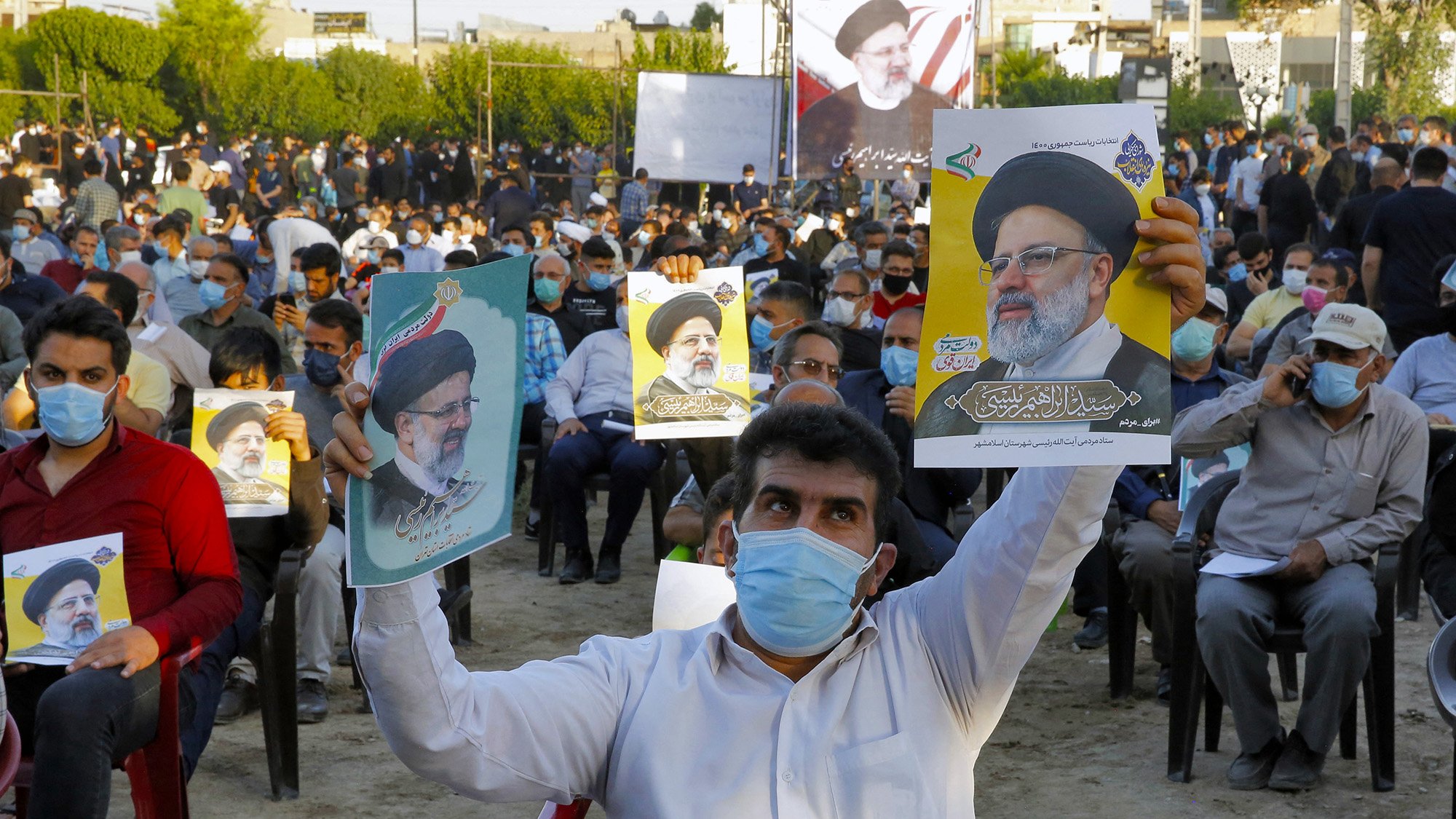 در تصویر، جمعیتی از افراد در یک فضای باز دیده میشود که روی صندلی نشستهاند و تعدادی از آنها در حال نگهداشتن پوسترهایی از یک شخصیت سیاسی هستند. مردم ماسک بهصورت دارند و در پسزمینه، تعدادی پرچم و تابلوهای نمایشی نیز وجود دارد. این جمعیت به نظر میرسد که در یک رویداد یا حمایت سیاسی شرکت کردهاند.