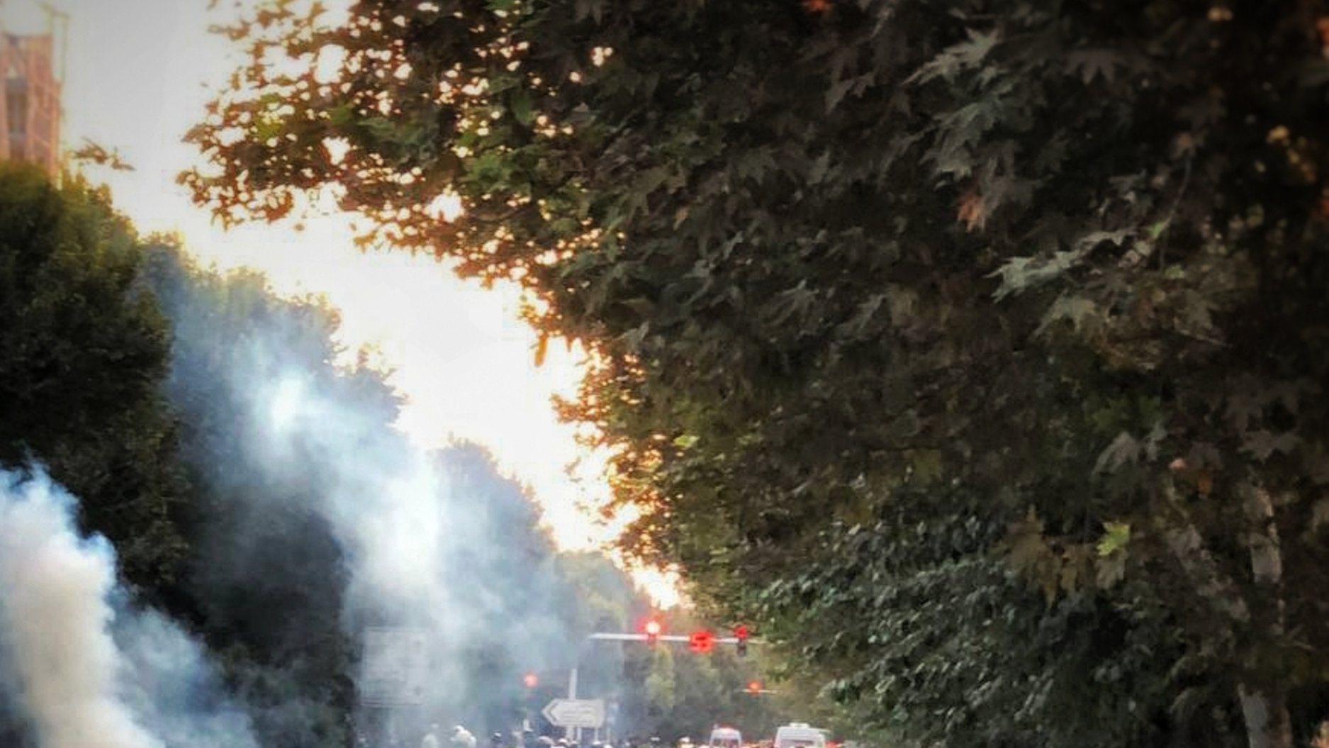 در این تصویر، صحنهای از یک شورش یا تظاهرات مشاهده میشود. فردی با پشت به دوربین در حال پرتاب شیئی به سمت جمعیت است. در اطراف او، دود غلیظی به هوا بلند شده و چند خودرو در نزدیکی محل حاضر هستند. درختان در دو سمت خیابان دیده میشوند و به نظر میرسد که فضای تنشزایی در حال ایجاد است. ترافیک و حضور مردم در صحنه نشاندهنده وضعیت پر سر و صدا و احتمالا ناآرام است.
