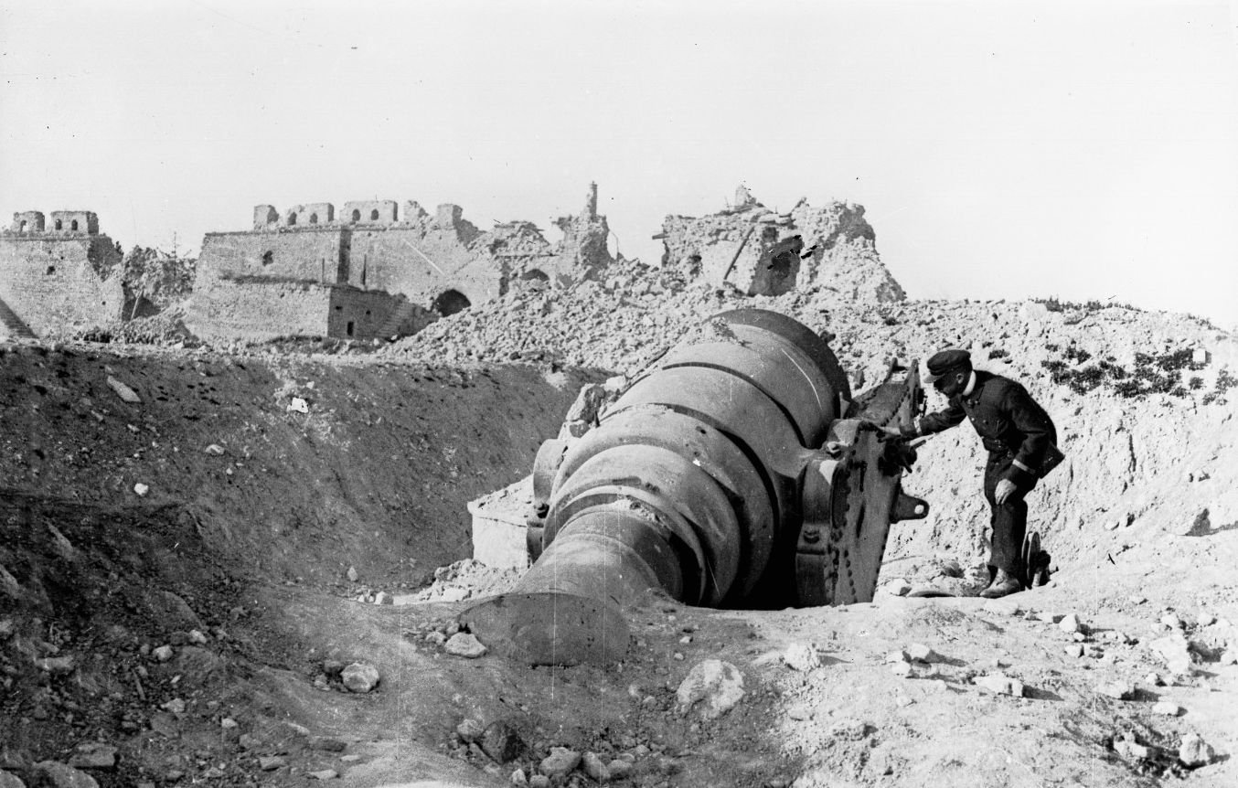 این تصویر به نظر میرسد نمایی تاریخی از یک مکان جنگی باشد. در تصویر، یک مرد به یک توپ بزرگ که به سمت زمین خوابیده است، نزدیک میشود. پسزمینه شامل ویرانههایی از ساختمانها یا قلعههای قدیمی است که گویای تخریب ناشی از جنگ یا نبردی است. همچنین، زمین اطراف آن سنگریزهها و خاکی دارد که نشاندهنده شرایط ناهموار و دشوار میباشد. به طور کلی، این تصویر حس غم و خرابی را منتقل میکند و یادآور هزینههای جنگ است.
