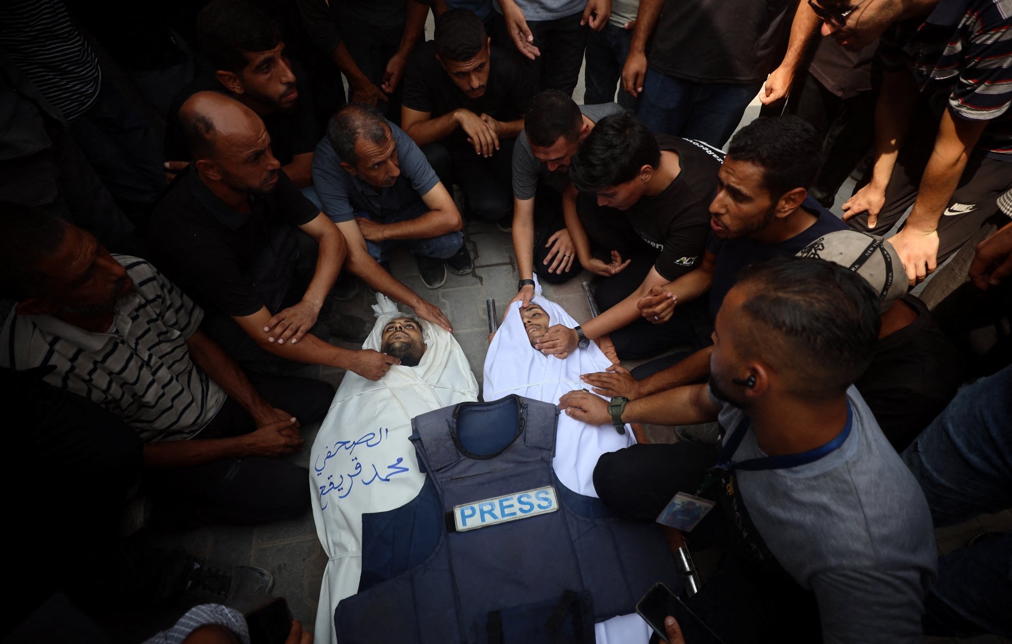 تصویری از گروهی از مردان که دور یک فرد دراز کشیده با پوششی به رنگ سفید و لباس خبرنگاری جمع شدهاند.