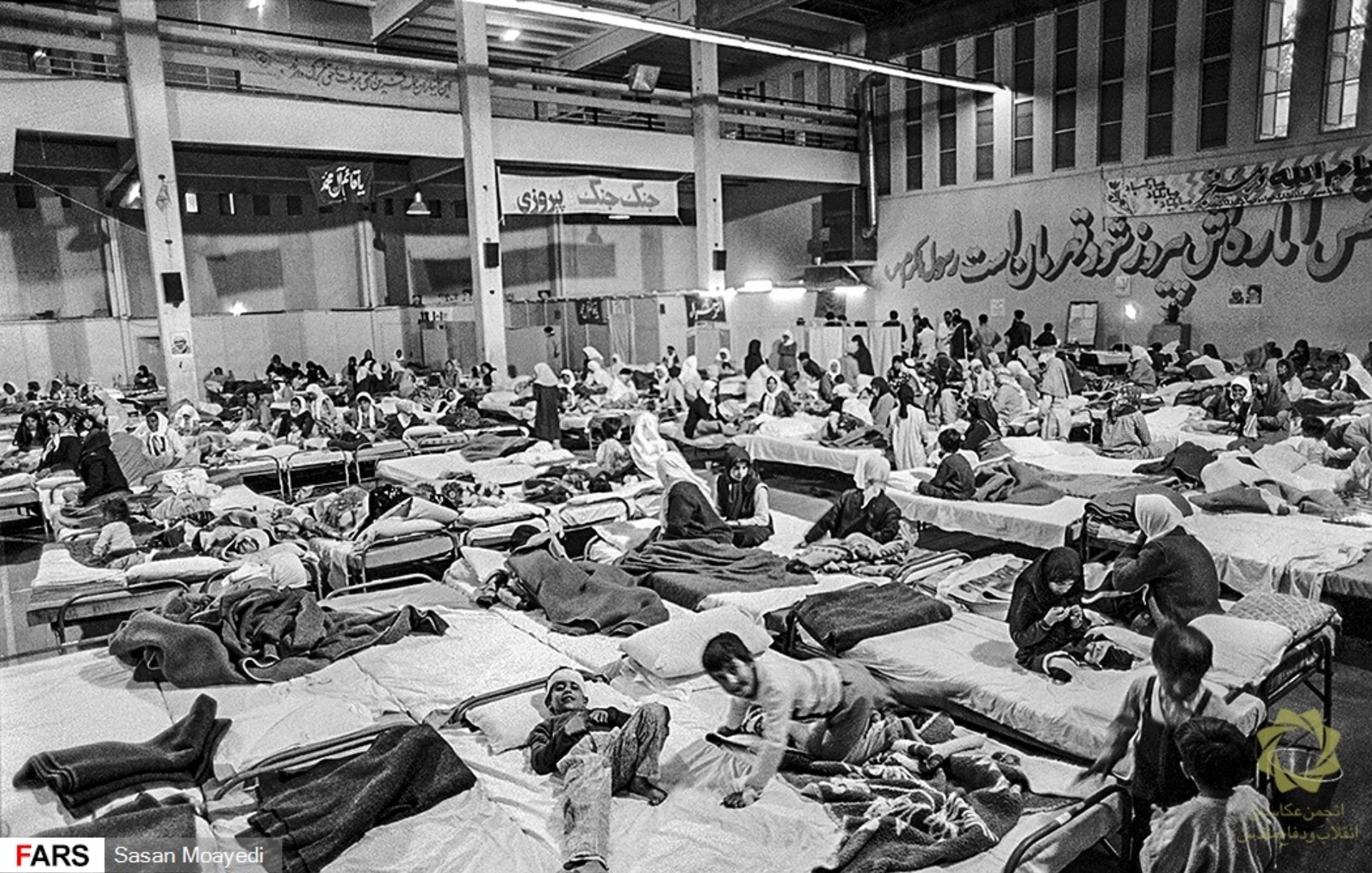 این تصویر یک فضای داخلی را نشان میدهد که پر از تختهای بیمارستانی است. افراد مختلفی در حالتهای مختلف دراز کشیدهاند، برخی در حال استراحت و برخی دیگر در حال حرکت یا صحبت با یکدیگر هستند. محیط به نظر شلوغ و پر از فعالیت میرسد، و پوشش بیشتر افراد ساده و شامل چادر یا پتو است. دیوارها به نوعی دکور شدهاند، و فضای کلی شاید به یک مرکز درمانی یا پناهگاه اشاره دارد.
