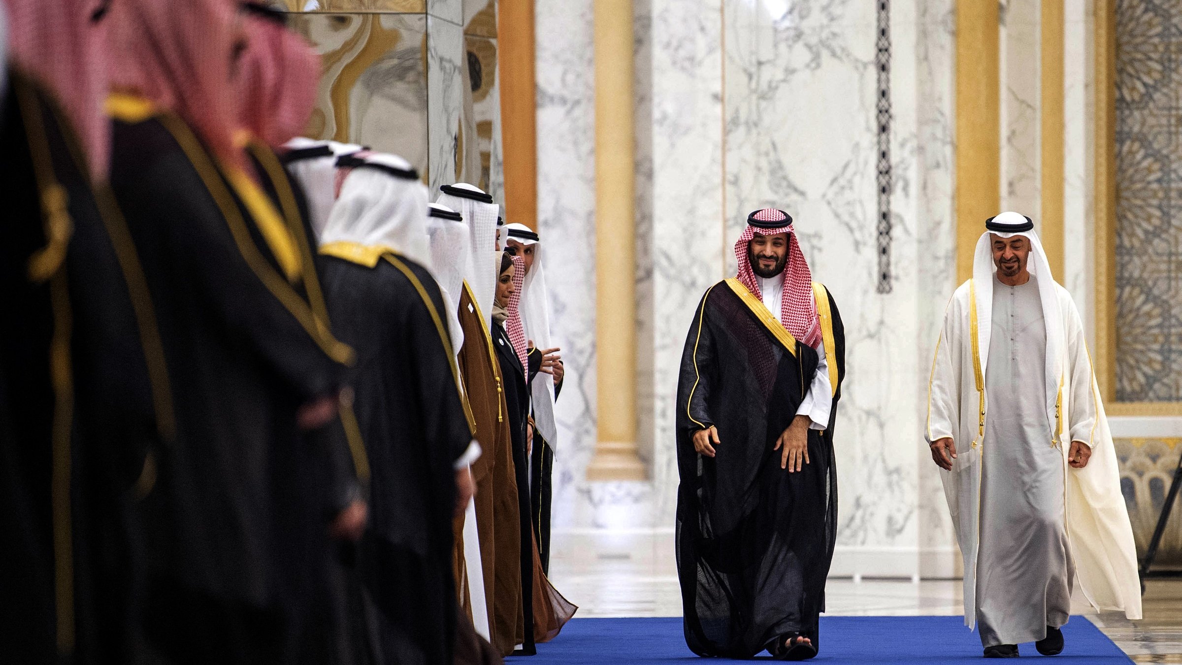 در این تصویر، گروهی از مردان به صورت رسمی در یک محیط داخل ساختمان قرار دارند. برخی از آنها لباسهای سنتی عربی به تن دارند و در حال خوشآمدگویی یا استقبال از یک نفر دیگر هستند که به آرامی در حال عبور از میان آنهاست. شخصی که در مرکز تصویر قرار دارد، لباسی متفاوت و برجستهتر دارد و به نظر میرسد نقش مهمتری در این مراسم بازی میکند. محیط با طراحی مجلل و دیوارهای سنگی تزئین شده است.