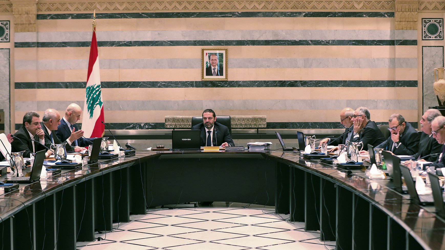 تصویر یک جلسه رسمی دولتی در لبنان را نشان میدهد. تعدادی از مقامات و اعضای دولت در اطراف یک میز بزرگ نشستهاند. در مرکز جلسه، یک مرد با لباسی رسمی و آرامش بر روی صندلی ریاست نشسته است و به دیگران نگاه میکند. در پسزمینه، پرچم لبنان و تصویری از یک شخصیت مهم بر دیوار نصب شده است. فضای اتاق با خطوط عمودی و افقی تزئینی و رنگهای ملایم طراحی شده است. افراد حاضر در جلسه به موضوعات مختلفی مشغول بحث و تبادل نظر هستند.