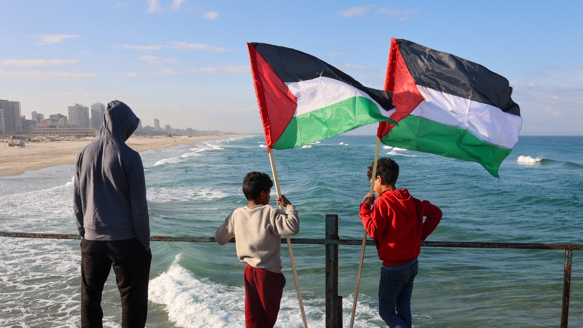 در این تصویر، سه کودک در کنار دریا ایستادهاند و به سمت امواج دریا نگاه میکنند. یکی از کودکان پرچم فلسطین را در دست دارد و دو نفر دیگر نیز در حال نظارت بر صحنه هستند. لباسهای آنها متنوع است و در پسزمینه میتوان نمایی از سواحل و ساختمانهای شهری را مشاهده کرد. آسمان آبی و ابرهای کمی در بالای تصویر وجود دارد که حال و هوای دلپذیری را به تصویر میدهد.