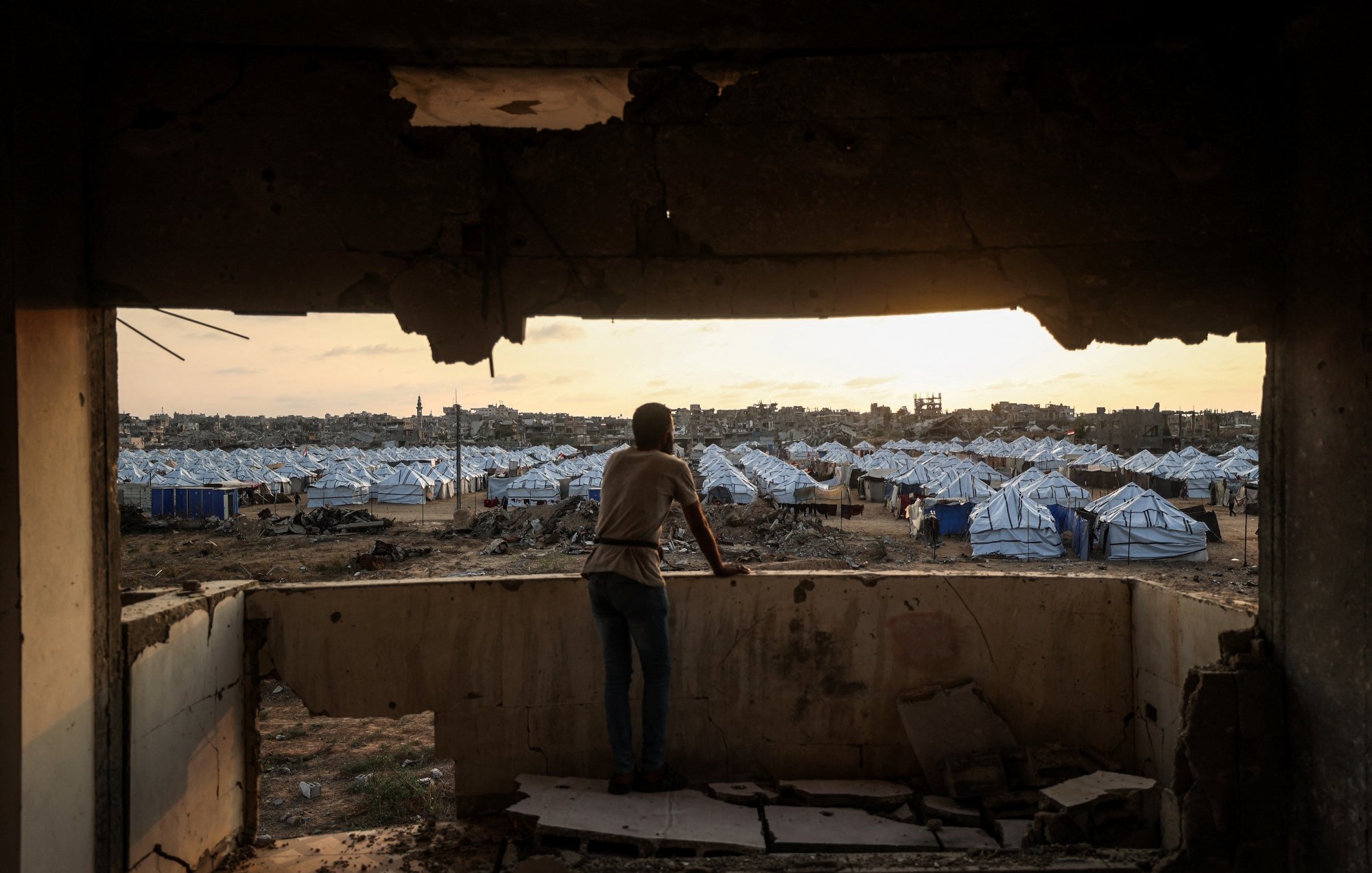 پسری از لبهی یک ساختمان ویران به سمت اردوگاه چادرها در غروب نگاه میکند.