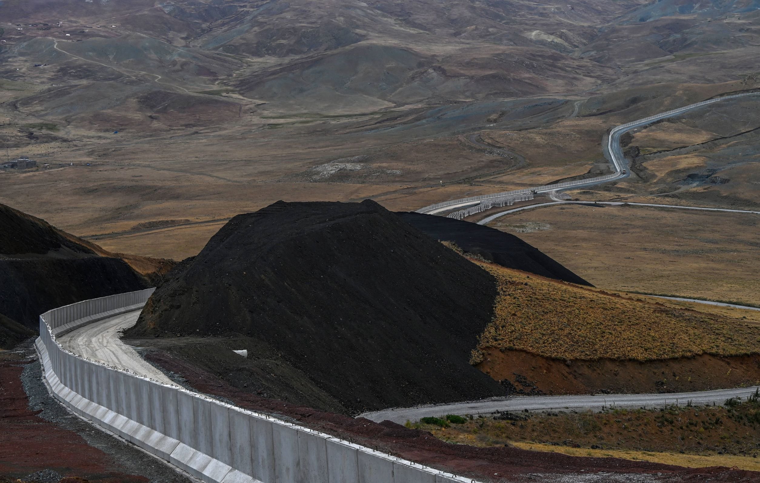 این تصویر نمایی از یک جاده در میان مناظر طبیعی خشک و کوهستانی را نشان میدهد. جاده به صورت منحنی و پیچدرپیچ از روی زمینهای ناهموار عبور میکند و در سمت راست تصویر، دیواری سفیدرنگ وجود دارد که به طور راستا در حال پیشروی است. در پسزمینه، تپهها و دامنههای کوهستانی با رنگهای ملایم و خاصی دیده میشوند که نمایی طبیعی و بکر را به تصویر میبخشند. فضای کلی تصویر به نوعی حس دورافتادگی و خلوتی را منتقل میکند.