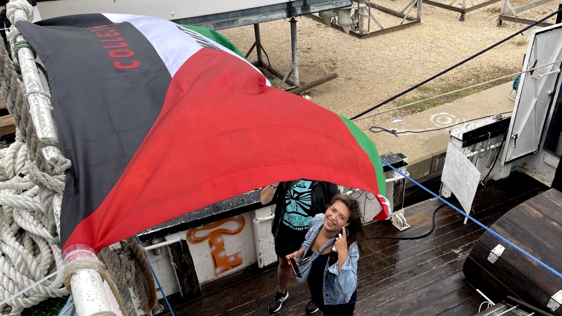 در این تصویر، دو نفر در حال کار با یک پرچم هستند که بر روی یک قایق یا سکوی چوبی قرار دارد. پرچم دارای رنگهای مشخصی است و یکی از افراد به دوربین لبخند میزند و انگشت شست خود را بالا برده است. محیط اطراف شامل تجهیزات قایقسواری و فضای باز است. به نظر میرسد این افراد در حال آمادهسازی یا نصب پرچم هستند.