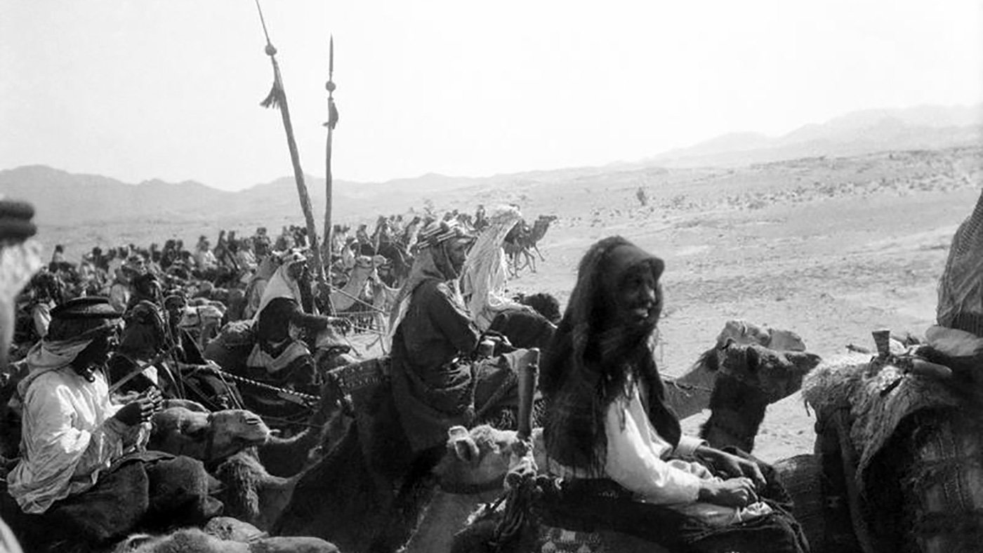 تصویری سیاه و سفید از یک کاروان در بیابان است. گروهی از افرادی که بر روی شترها سوار هستند، در حال حرکت به سمت دوربین هستند. در میان آنها زنانی با پوششهای سنتی و مردانی با لباسهای مختلف دیده میشوند. برخی از افراد نیز پرچمهایی در دست دارند. پسزمینه تصویر شامل تپههای شنی و کوهها است که حس فضای بیابانی را القا میکند. جو تصویر قدیمی و تاریخی به نظر میرسد.