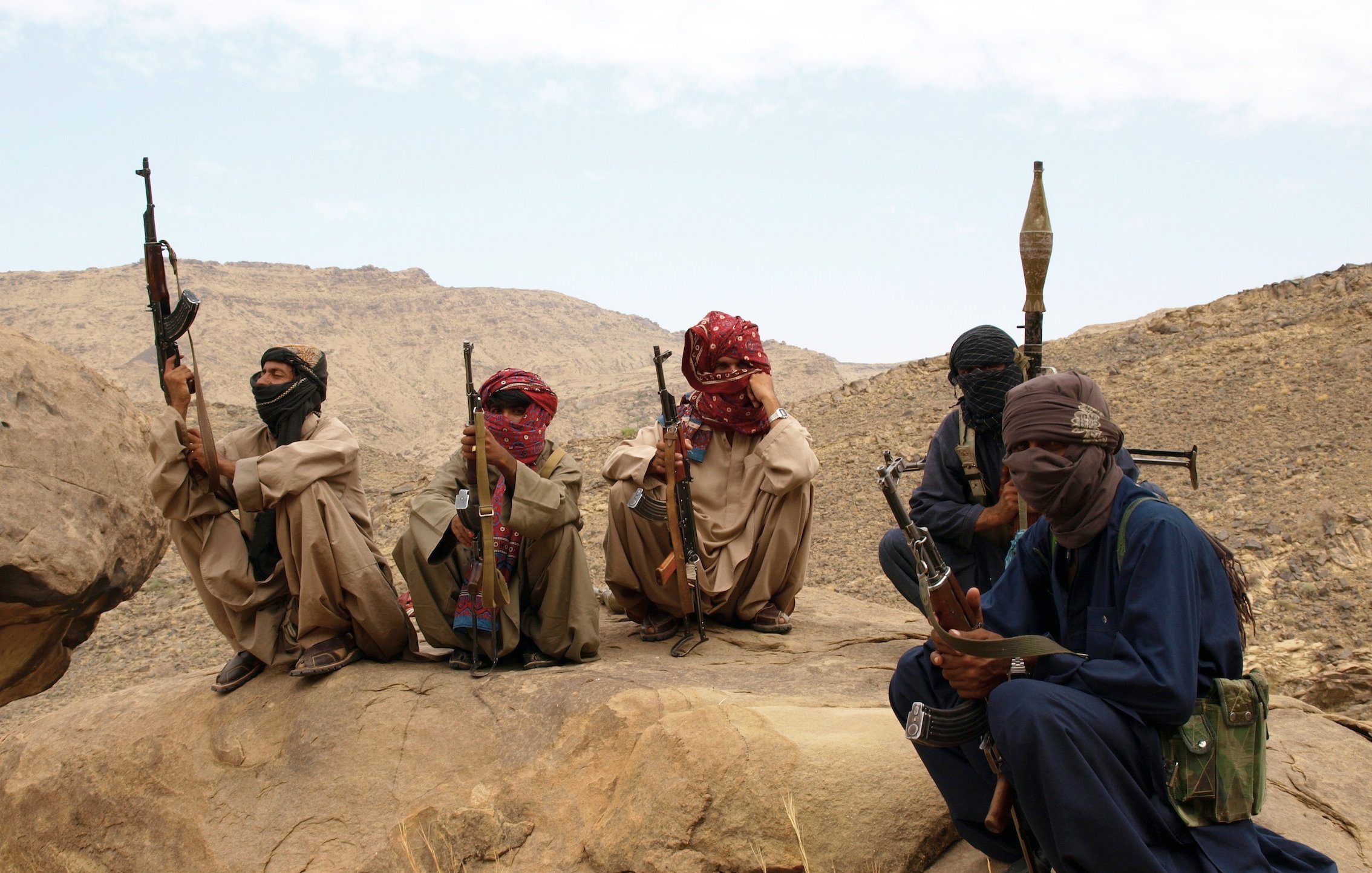 تصویر گروهی از مردان مسلح را نمایش میدهد که در یک منطقه بیابانی یا کوهستانی نشستهاند. آنها لباسهای سنتی محلی به تن دارند و صورتهایشان با پوشاکی مثل شال یا دستمال پوشانده شده است. برخی از آنها تفنگها و سلاحهای دیگری در دست دارند، در حالی که در حال نظارت بر محیط اطراف خود هستند. پسزمینه شامل سنگها و تپههای بیابانی است. وضعیت عمومی تصویر حس جدیت و آمادگی را منتقل میکند.