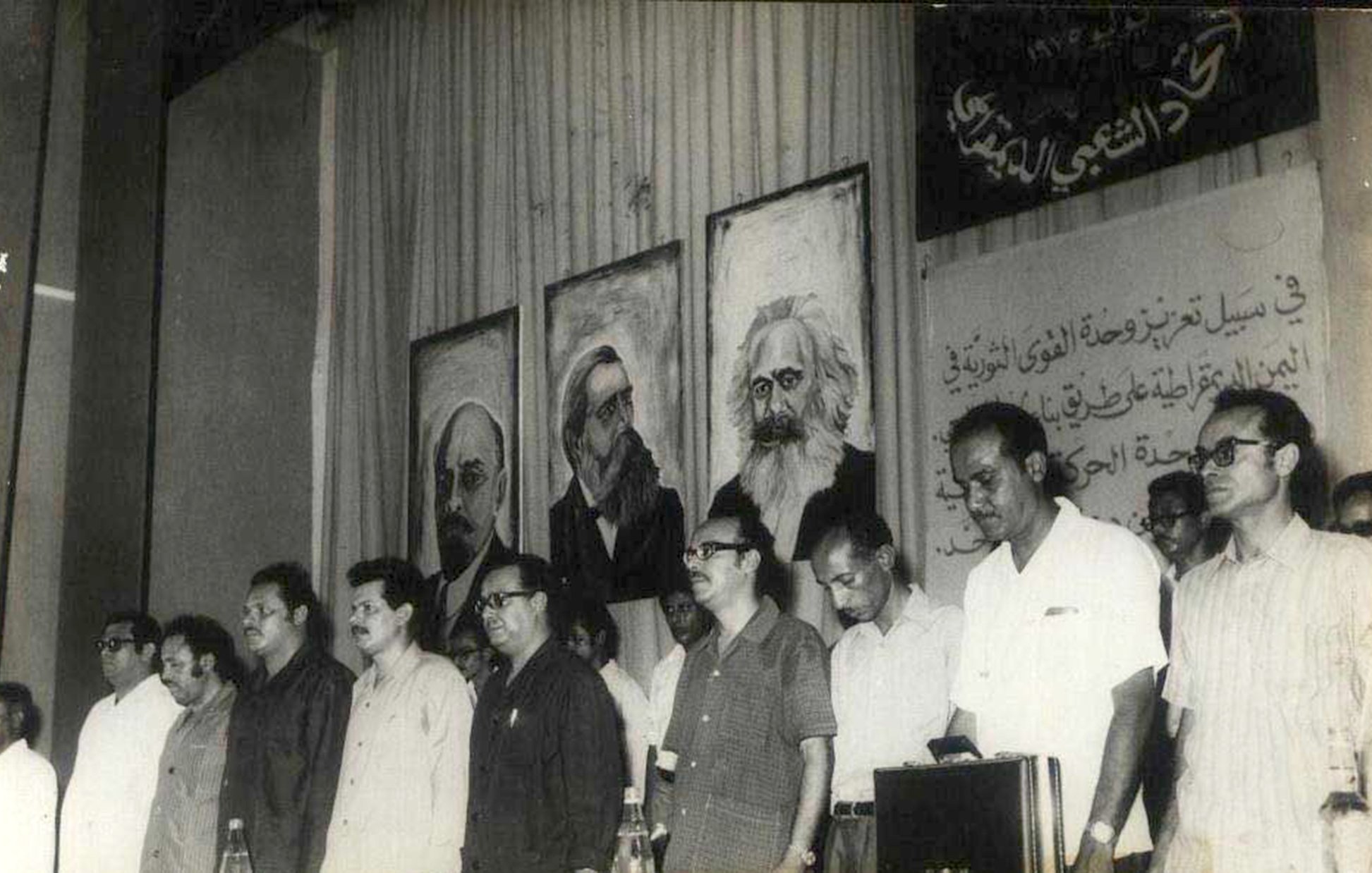 این تصویر مربوط به یک تجمع یا کنفرانس سیاسی به نظر میرسد. در پسزمینه، تابلوهایی از چهرههای معروف تاریخی یا سیاسی وجود دارد. افرادی که در جلوی صحنه ایستادهاند، موضع جدی و متفکرانهای دارند و در حال گوش دادن به سخنرانی یا برنامهای هستند. فضای کلی تصویر به نظر میرسد که تمرکز بر مسائل اجتماعی و سیاسی داشته باشد. دیوار پشتشان نیز با متنی به زبان عربی تزئین شده است.