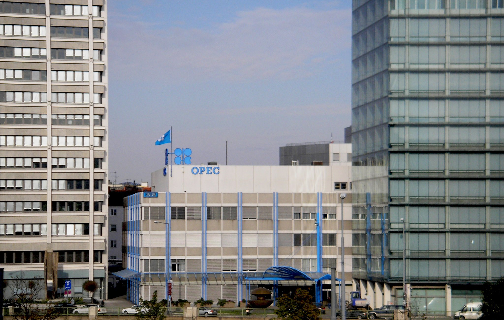 این تصویر نمایی از ساختمان اوپک (سازمان کشورهای صادرکننده نفت) را نشان میدهد. ساختمان به صورت مدرن و با طراحی سادهای ساخته شده و رنگ اصلی آن سفید و آبی است. در بالای ساختمان پرچم اوپک را میبینیم که به اهتزاز درآمده. در اطراف ساختمان، ساختمانهای بلند و مدرن دیگری نیز وجود دارند که فضای شهری را شکل میدهند. آسمان نیز کمی ابری به نظر میرسد.