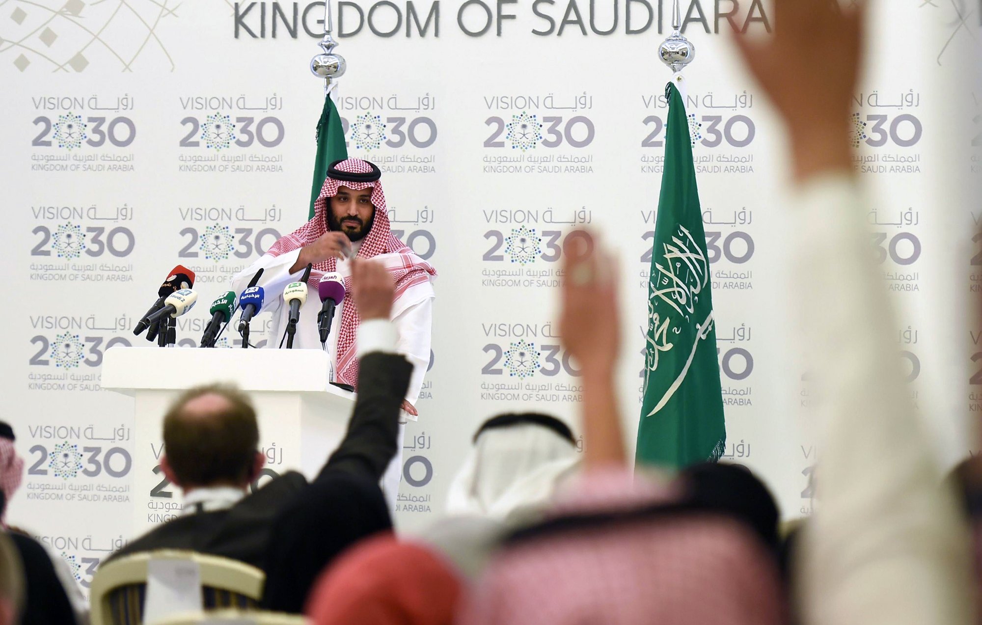 در این تصویر، یک مرد جوان با لباس سنتی سعودی و عمامه، در حال سخنرانی در مقابل میکروفنها و دوربینهای خبری است. او در پسزمینهای شامل پرچمهای عربستان سعودی و شعارهایی درباره "چشمانداز ۲۰۳۰" قرار دارد. تعدادی از افراد نشسته در سالن، دستهای خود را برای پرسش بالا بردهاند. فضا به وضوح نشاندهنده یک کنفرانس خبری یا رویداد رسمی است.