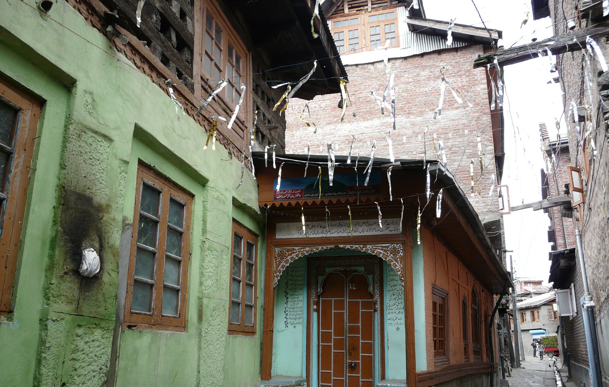 این تصویر نشاندهنده یک خیابان باریک در یک منطقه قدیمی است. ساختمانها به سبک سنتی ساخته شدهاند و رنگهای مختلفی دارند. دیوار یک ساختمان به رنگ سبز و دیگری به رنگ نارنجی است که با جزئیات چوبی تزئین شده است. درب چوبی و پنجرههای با قاب چوبی، زیبایی خاصی به این فضا بخشیدهاند. درختان و لامپهای آویزان نیز جزو دیگر عناصر موجود در تصویر هستند که به فضایی دلانگیز و سنتی افزودهاند.