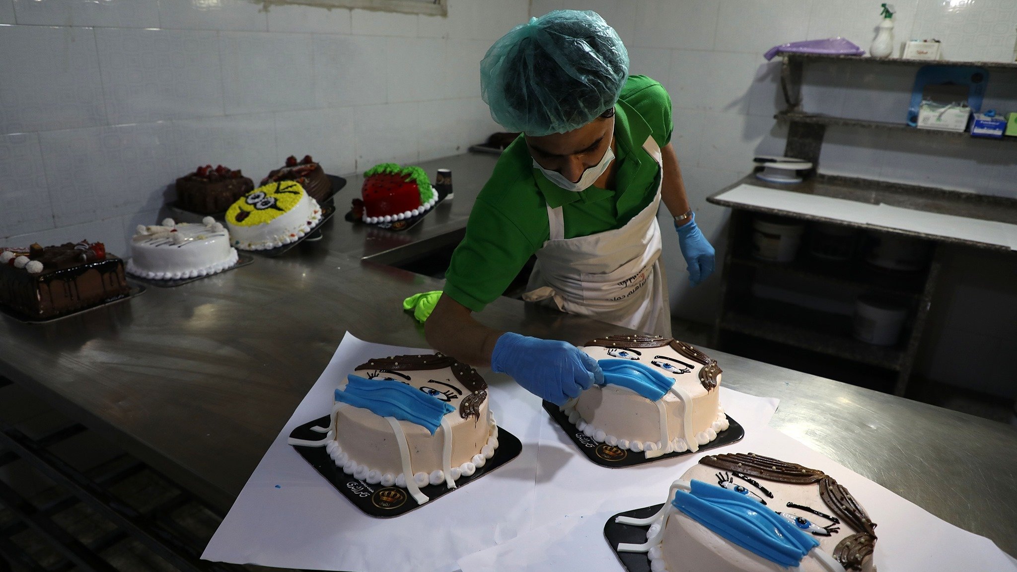 در تصویر یک نانوایی یا قنادی دیده میشود که یک کارگر در حال تزئین کیکهاست. این کیکها شکل و طراحی خاصی دارند و بر روی آنها معمولاً تصاویر چهرهای با ماسک قرار داده شده است. کارگر با دقت کیکها را تزئین میکند و از دستکش و کلاه مخصوص استفاده میکند. در پسزمینه، تعدادی کیک دیگر نیز قرار دارند که آماده فروش هستند. فضای قنادی تمیز و مرتب به نظر میرسد.