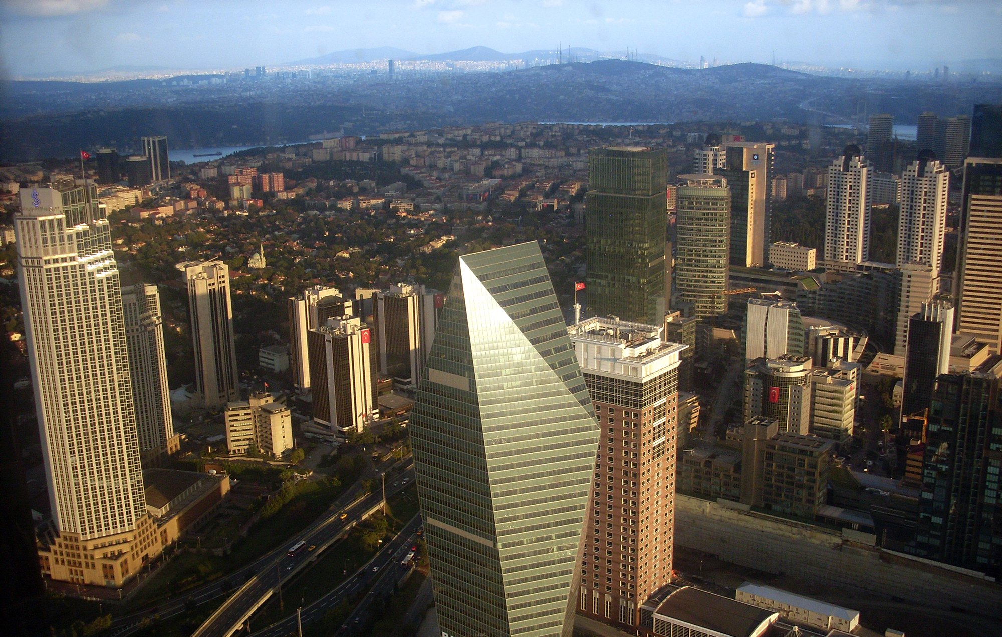 این تصویر نمایی از یک شهر مدرن است که در آن ساختمانهای بلند و شیک مشاهده میشوند. در مرکز تصویر یک ساختمان با طراحی متمایز و زاویهدار وجود دارد که بهنظر میرسد نماد یکی از بانکها باشد. دیگر ساختمانها نیز به ارتفاع و تنوع طراحی در اطراف آن قرار دارند. فضای سبز و خیابانهای در حال تردد نیز در بخشهای پایین تصویر قابل مشاهده است. آسمان آبی با ابرهای خفیف به زیبایی تصویر افزوده است.