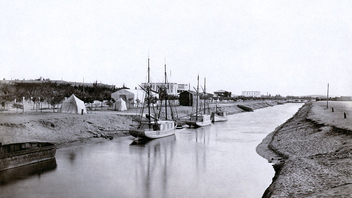 تصویر نمایی قدیمی از یک رودخانه یا کانال است که در دو طرف آن قایقها، به ویژه قایقهای بادبانی، قرار دارند. در سمت راست تصویر، چندین قایق به آرامی در آب شناور هستند و در سمت چپ، بخشی از ساحل با شن و ماسه دیده میشود. در پسزمینه، چادرها و ساختمانهایی به چشم میخورد که نشاندهنده حضور انسانی و فعالیتهای مربوط به کشاورزی یا صید هستند. نور تصویر به نظر میرسد نرم و ملایم باشد که حس آرامشی را ایجاد میکند.