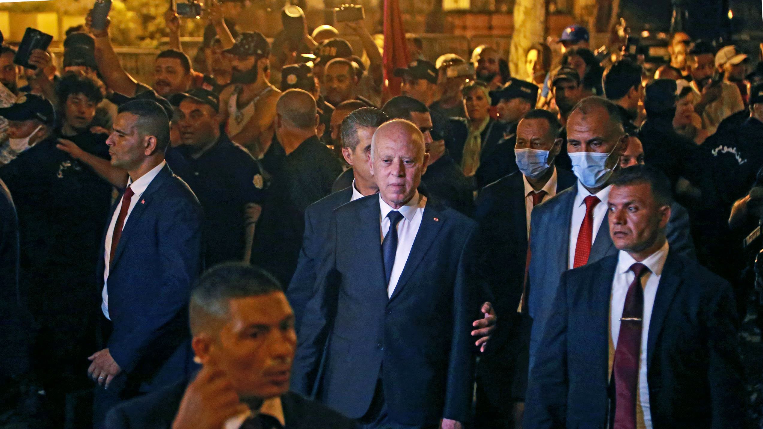 در این تصویر، گروهی از افراد در یک محیط شلوغ و پرجمعیت بهنظر میرسند. در مرکز تصویر، فردی با کت و شلوار تیره در حال عبور است و چندین محافظ یا همراه در اطراف او حضور دارند. برخی از افراد در زمینه تصویر، ماسک بهصورت دارند و دیگران مشغول تماشا و عکاسی از صحنه هستند. محیط به نظر میرسد شبانه باشد و نورها بهخوبی در تصویر مشاهده میشوند.