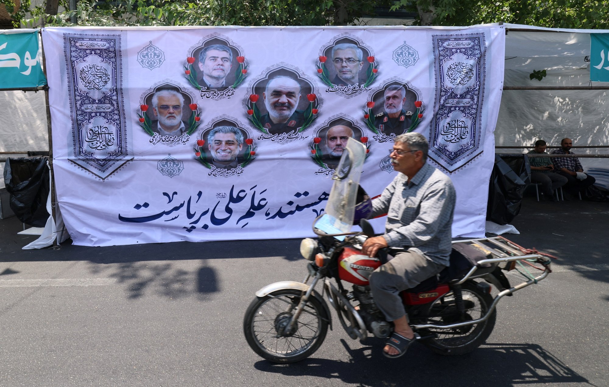 تصویر مردی سوار بر موتورسیکلت که در پیشزمینه، بنر بزرگ و مردم نشسته در پشت آن دیده میشوند.