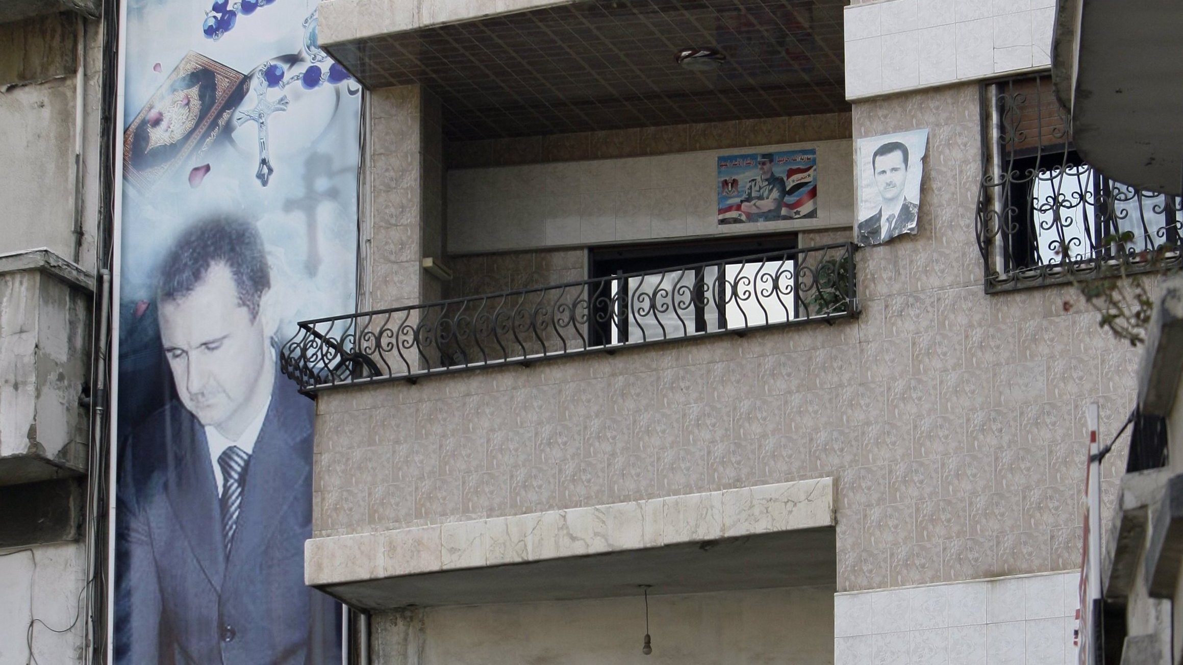 در این تصویر، نمایی از یک ساختمان چند طبقه دیده میشود که در آن پوسترها و تصاویر بزرگی از یک شخص (که احتمالاً یک شخصیت سیاسی است) نصب شده است. دو مرد در بالکن طبقه دوم ایستادهاند و به فضای بیرون نگاه میکنند. همچنین، در زیر ساختمان، بنرهایی با شعارها و تصاویری از دیگر افراد نصب شده است. محیط به نظر میرسد که در یک منطقه شهری باشد و برخی نشانههای ملی مانند پرچم در تصویر دیده میشود.