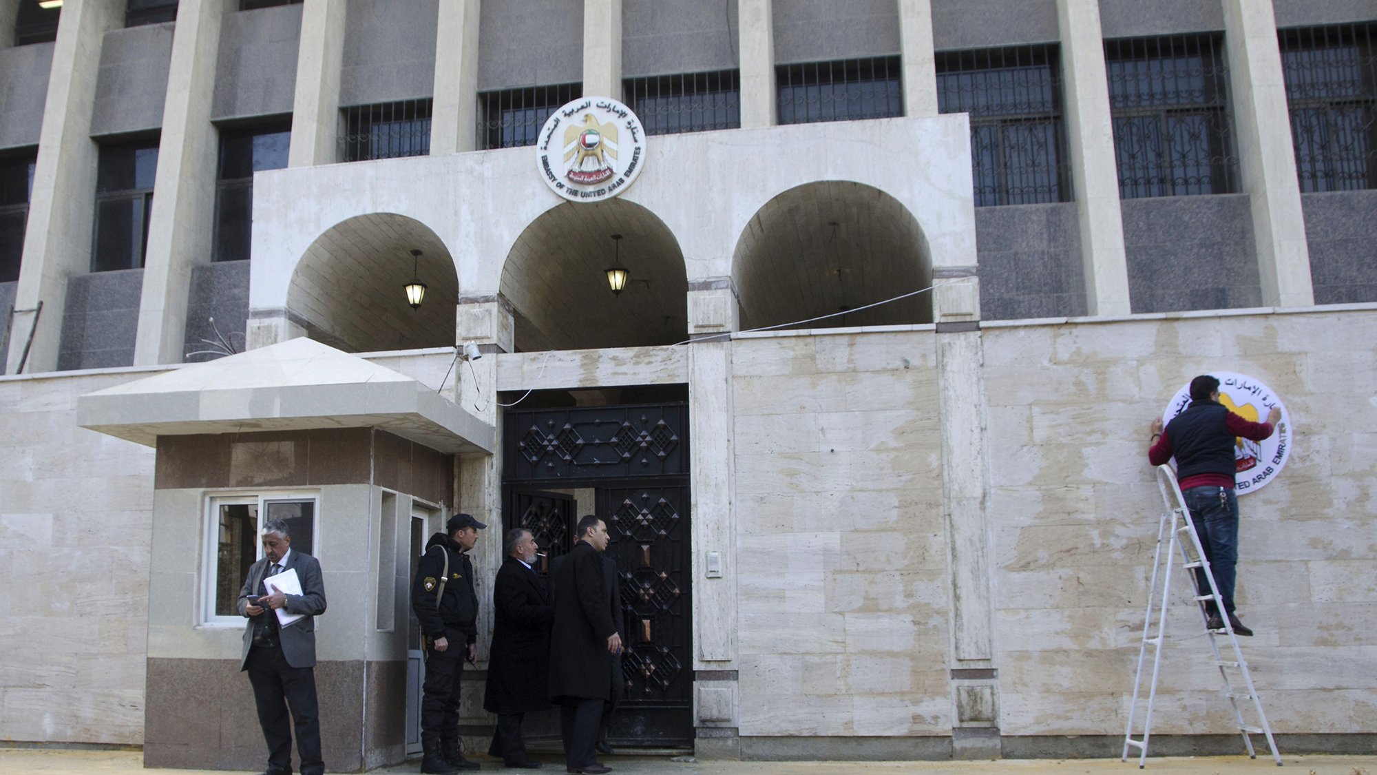 تصویر نشاندهندهٔ یک ساختمان دولتی است که به نظر میرسد در یک کشور عربی قرار دارد. در جلوی ساختمان، شبکهای از ستونها و پنجرهها وجود دارد و بر روی دیوار ورودی، نشان یا علامت مشخصی نصب شده است. چند نفر در جلوی ساختمان ایستادهاند؛ یکی در حال نصب علامت بر روی دیوار و دو نفر دیگر در حال گفتگو به نظر میرسند. همچنین، یک نگهبان در کنار ورودی قابل مشاهده است. زمینهٔ تصویر نیز آسفالت و خطکشی خیابان است.